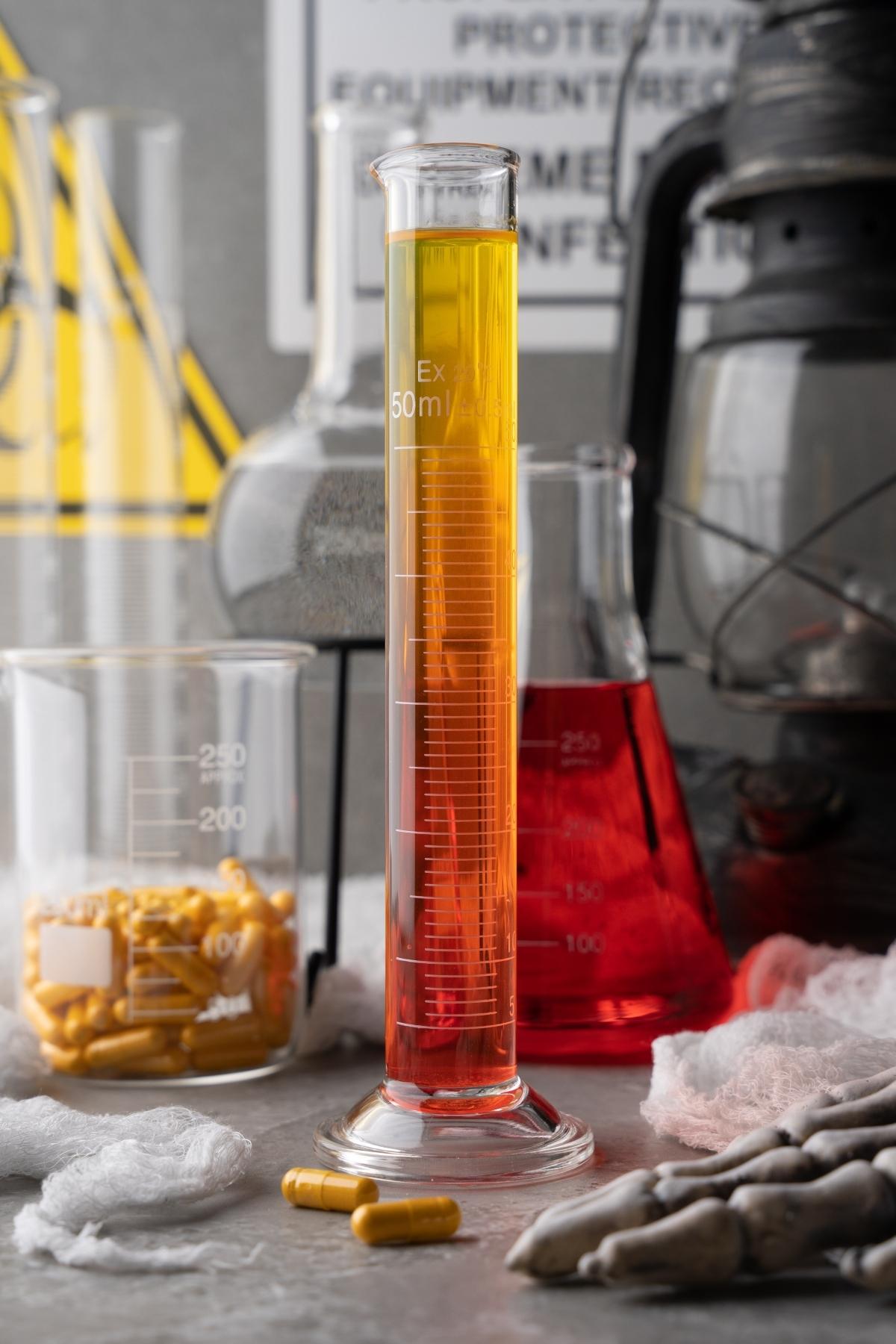 A graduated cylinder with orange liquid in a lab, surrounded by beakers, capsules, and lab equipment—perfect for experimenting with vitamins that glow under black light.