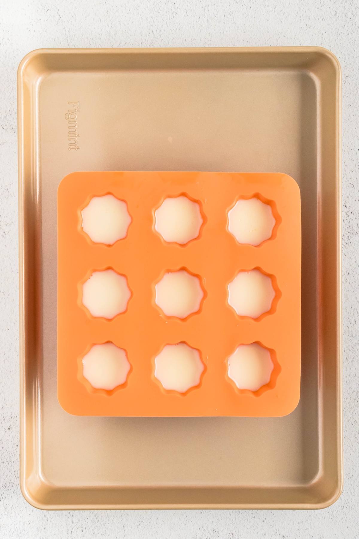 An orange silicone mold with eight filled cavities sits on a gold baking sheet.