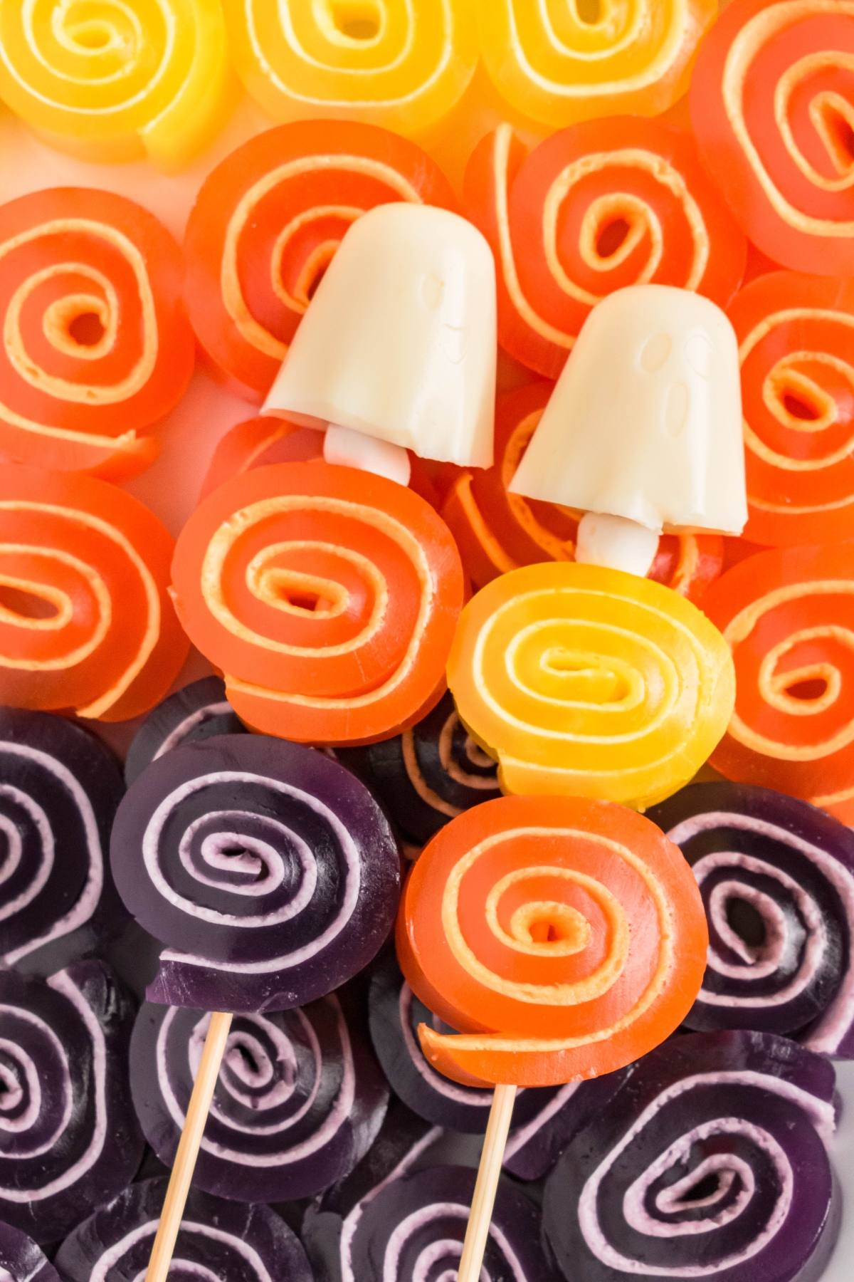 Colorful spiral gummy candies on sticks, with two white ghost-shaped candies, arranged in rows.