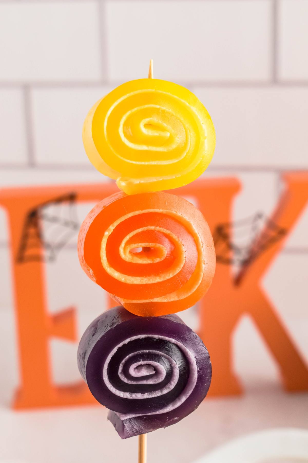Three rolled fruit leathers—yellow, orange, and purple—stacked on a skewer, with blurred letters in background.