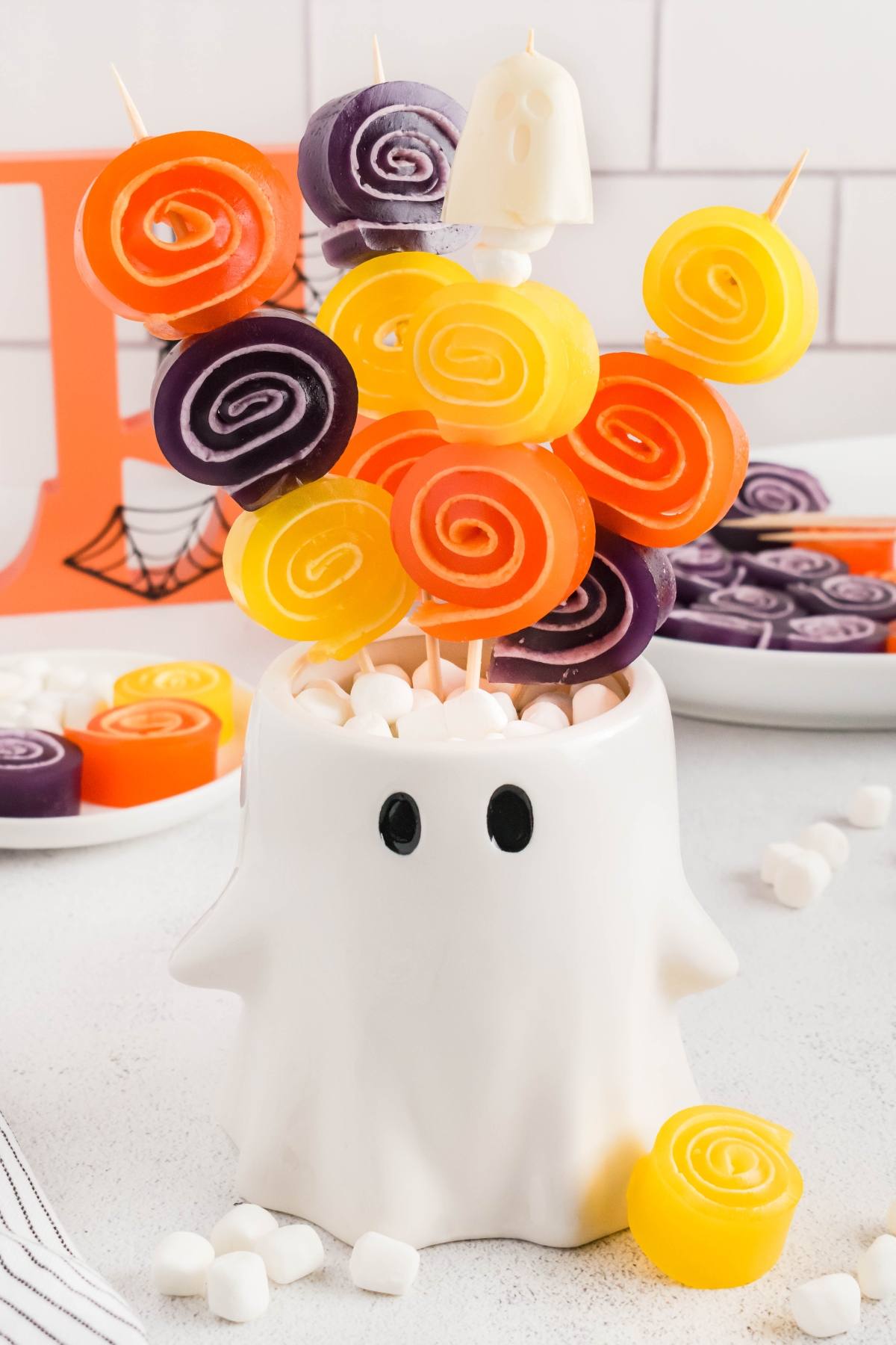Colorful swirl candies on skewers arranged in a white ghost-shaped holder with marshmallows.