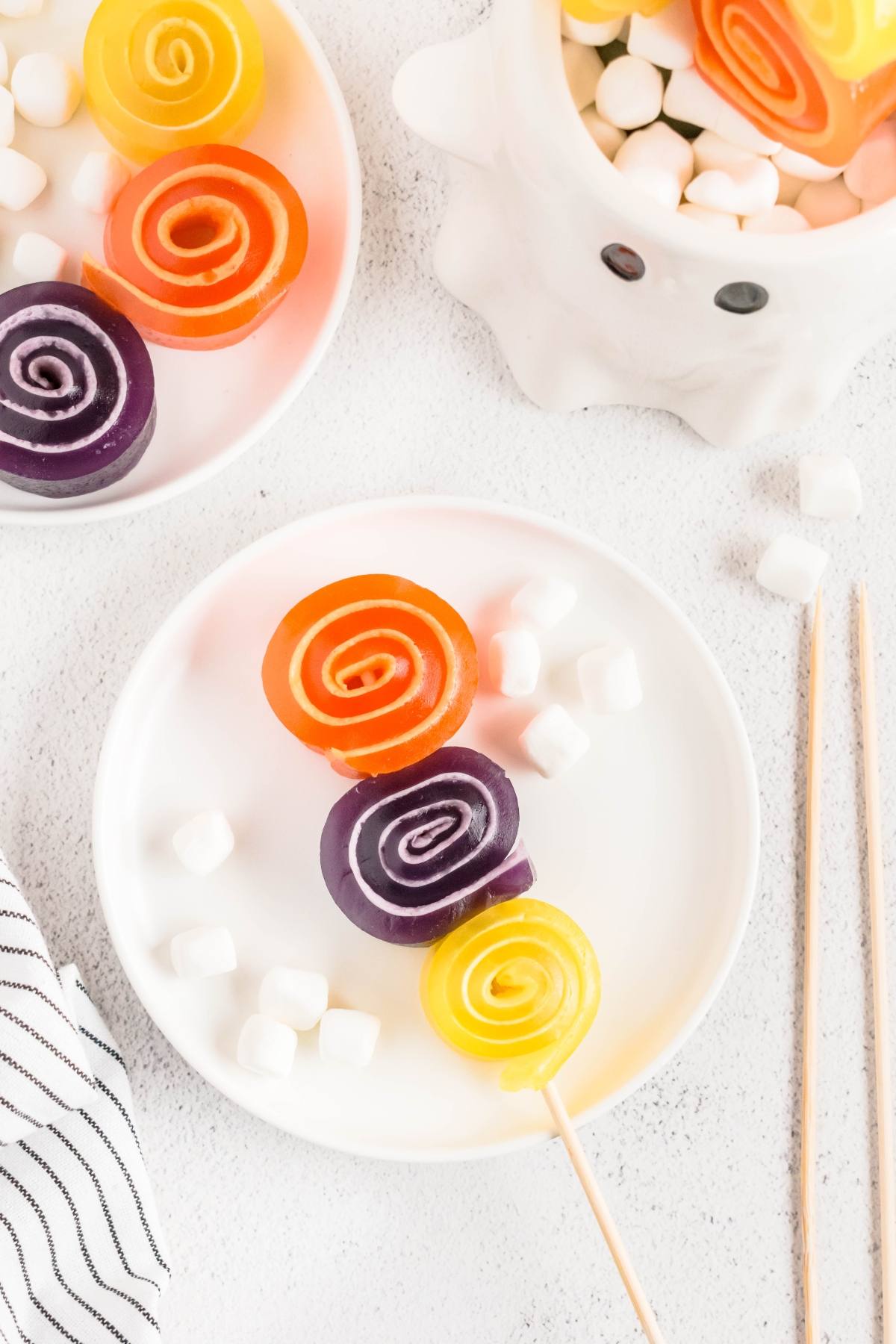 Colorful spiral gummy candies on skewers with mini marshmallows, served on white plates and a ghost-shaped bowl.