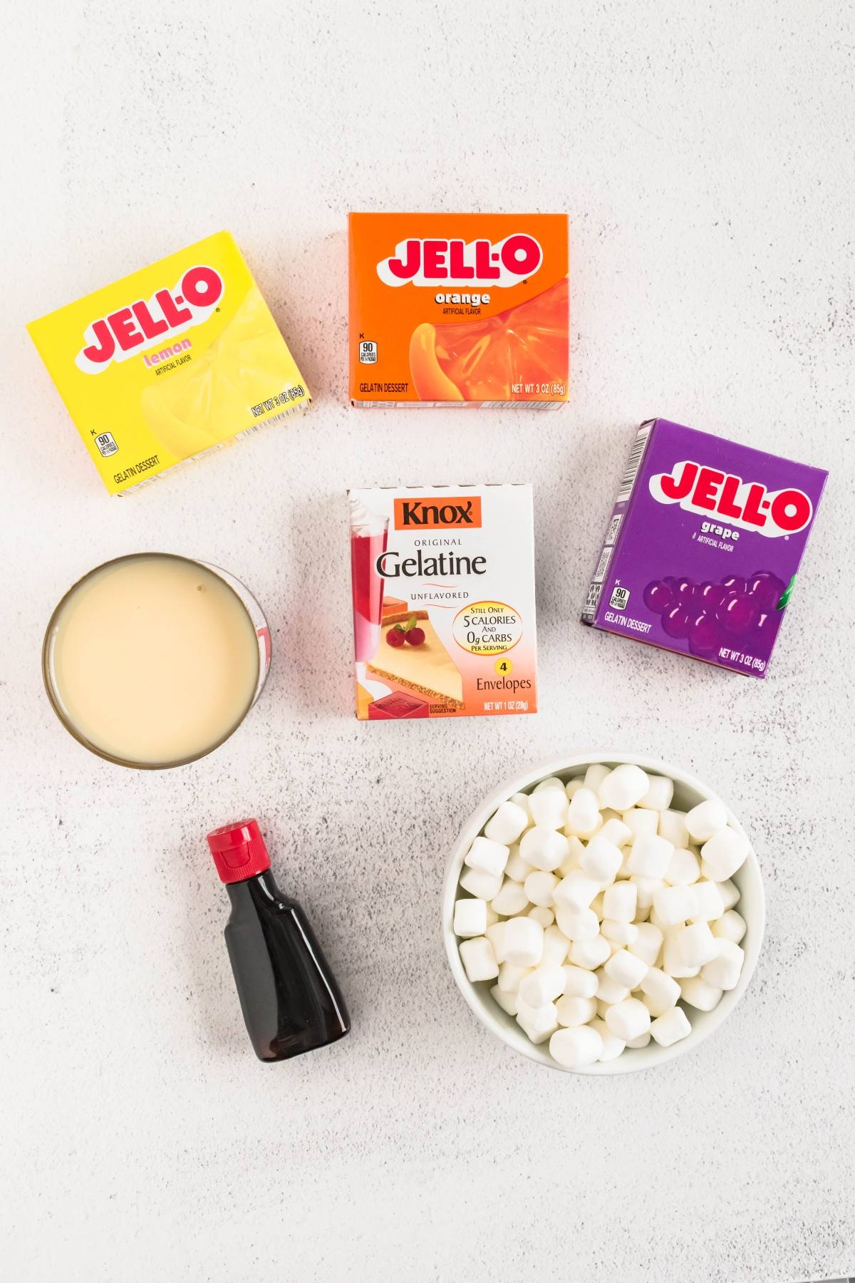 Boxes of lemon, orange, and grape Jell-O, gelatin, marshmallows, condensed milk, and vanilla on a white surface.