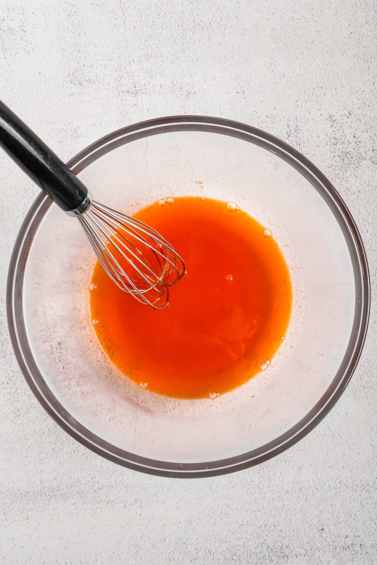 A glass bowl with orange liquid and a metal whisk on a light surface.