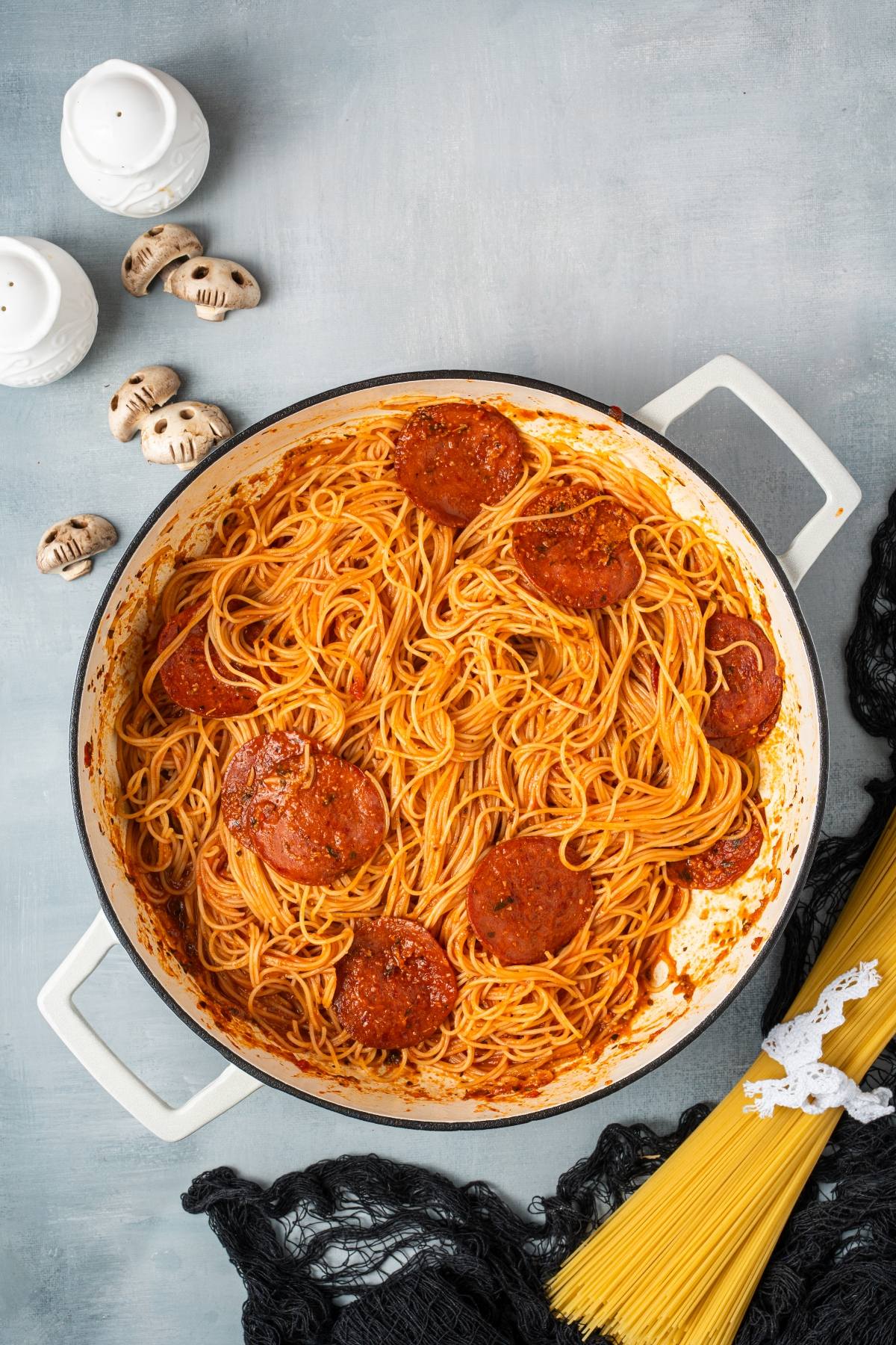 Pot of spaghetti with red sauce and round meat slices, surrounded by dry pasta and mushroom decorations.