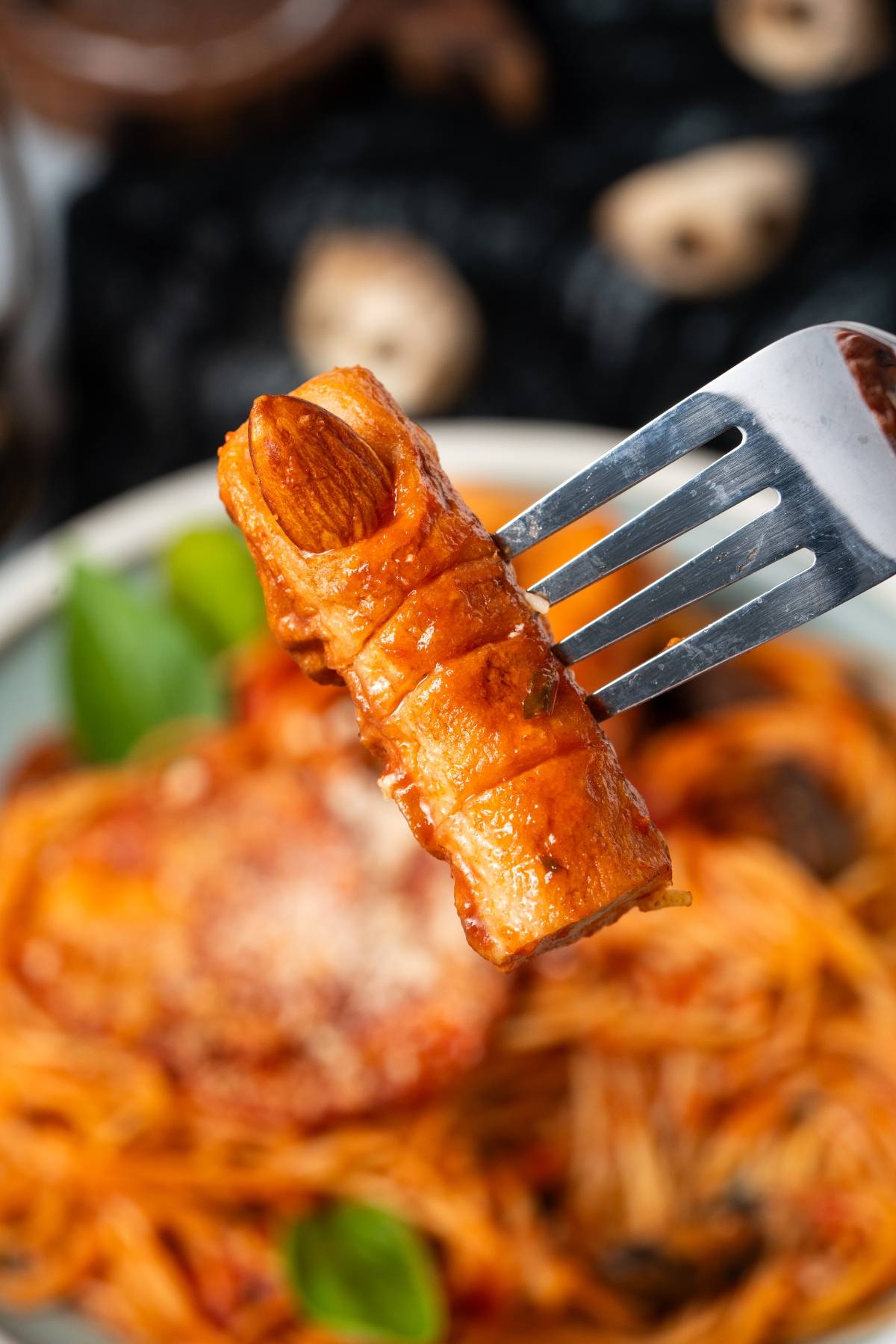 A fork holds a sausage made to look like a finger, with pasta and sauce in the background.