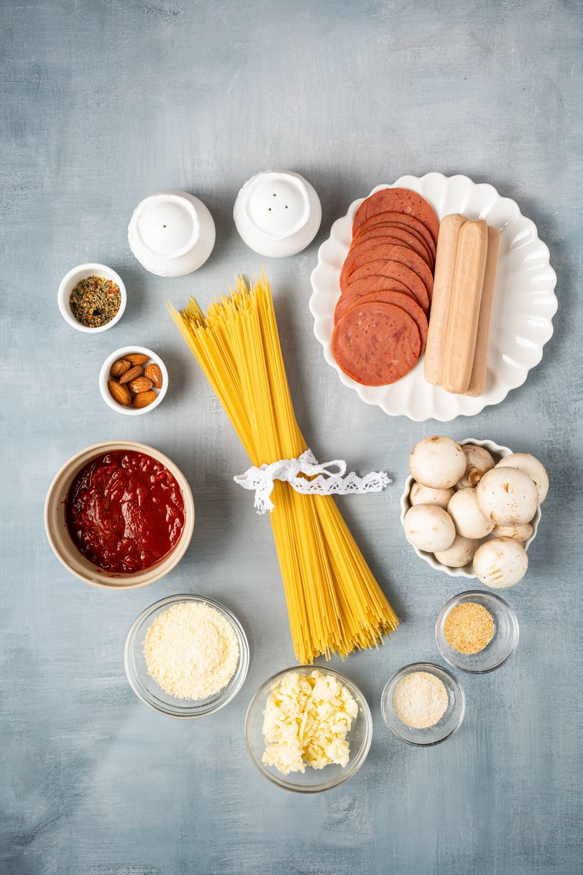 Uncooked spaghetti, mushrooms, tomato sauce, cheese, sausage, spices, and seasonings on a gray surface.