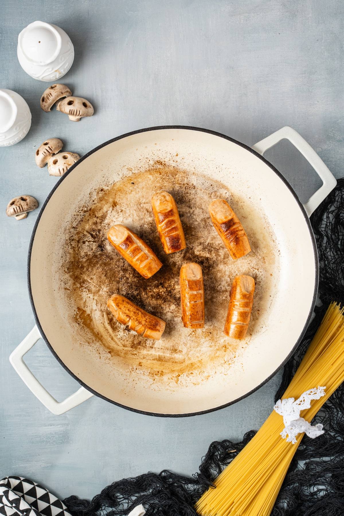 Six browned sausages cooking in a white pan beside uncooked spaghetti and mushrooms on a gray surface.