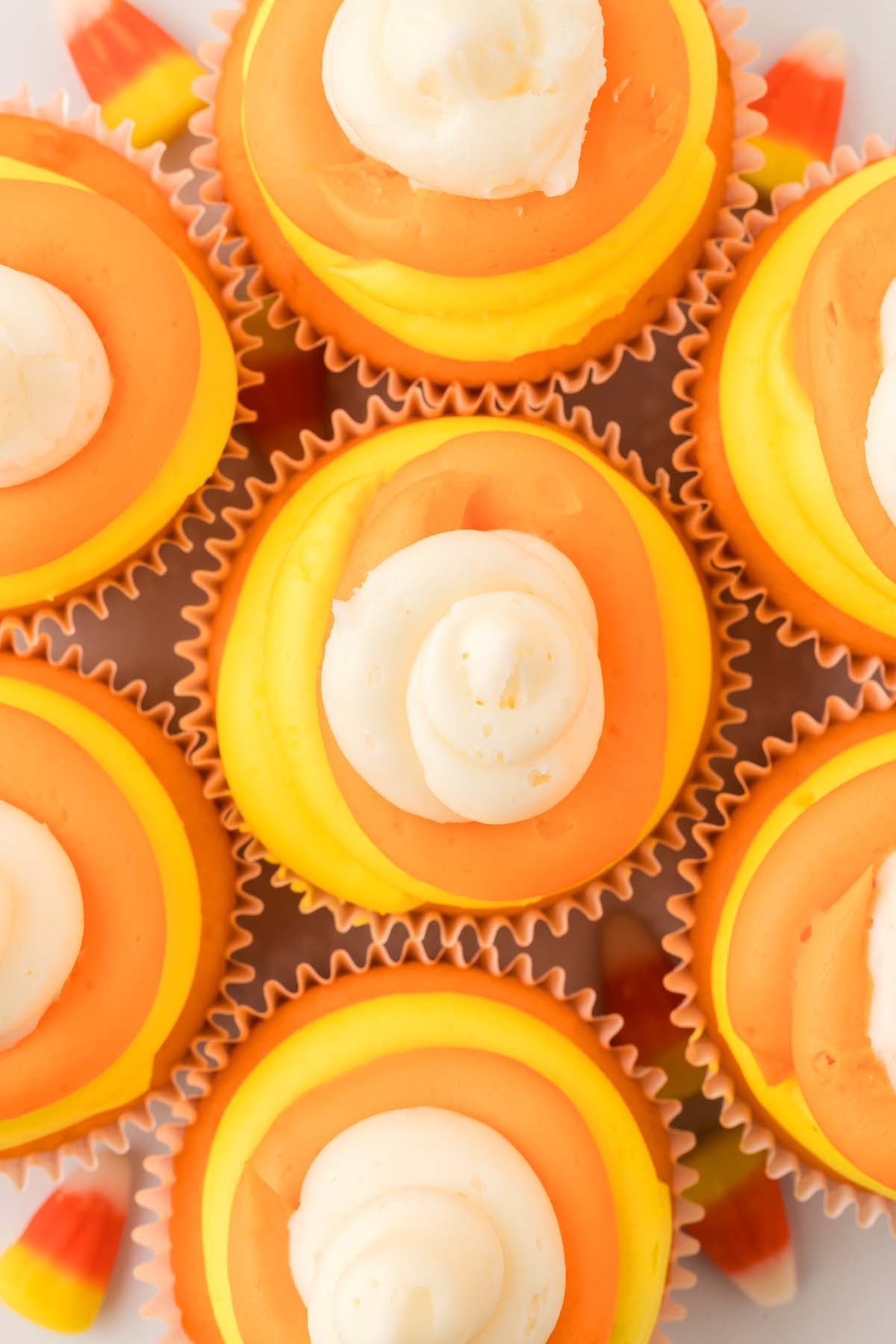 Six candy corn cupcakes with yellow, orange, and white frosting are arranged closely together, surrounded by festive candy corn pieces.