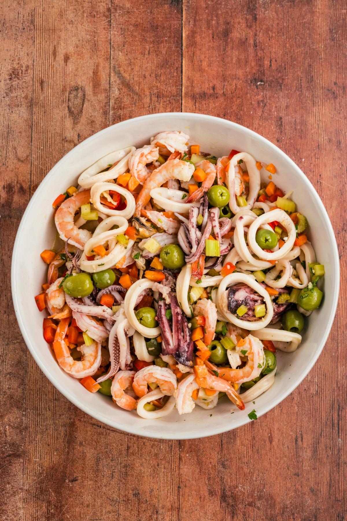Seafood salad with shrimp, calamari rings, olives, and chopped vegetables in a white bowl on a wooden table.