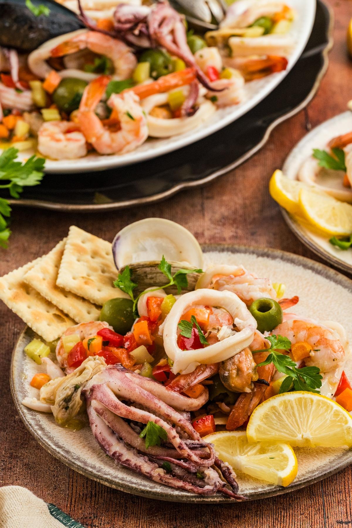 Seafood salad with shrimp, squid, olives, and vegetables, served with lemon wedges and crackers on a plate.