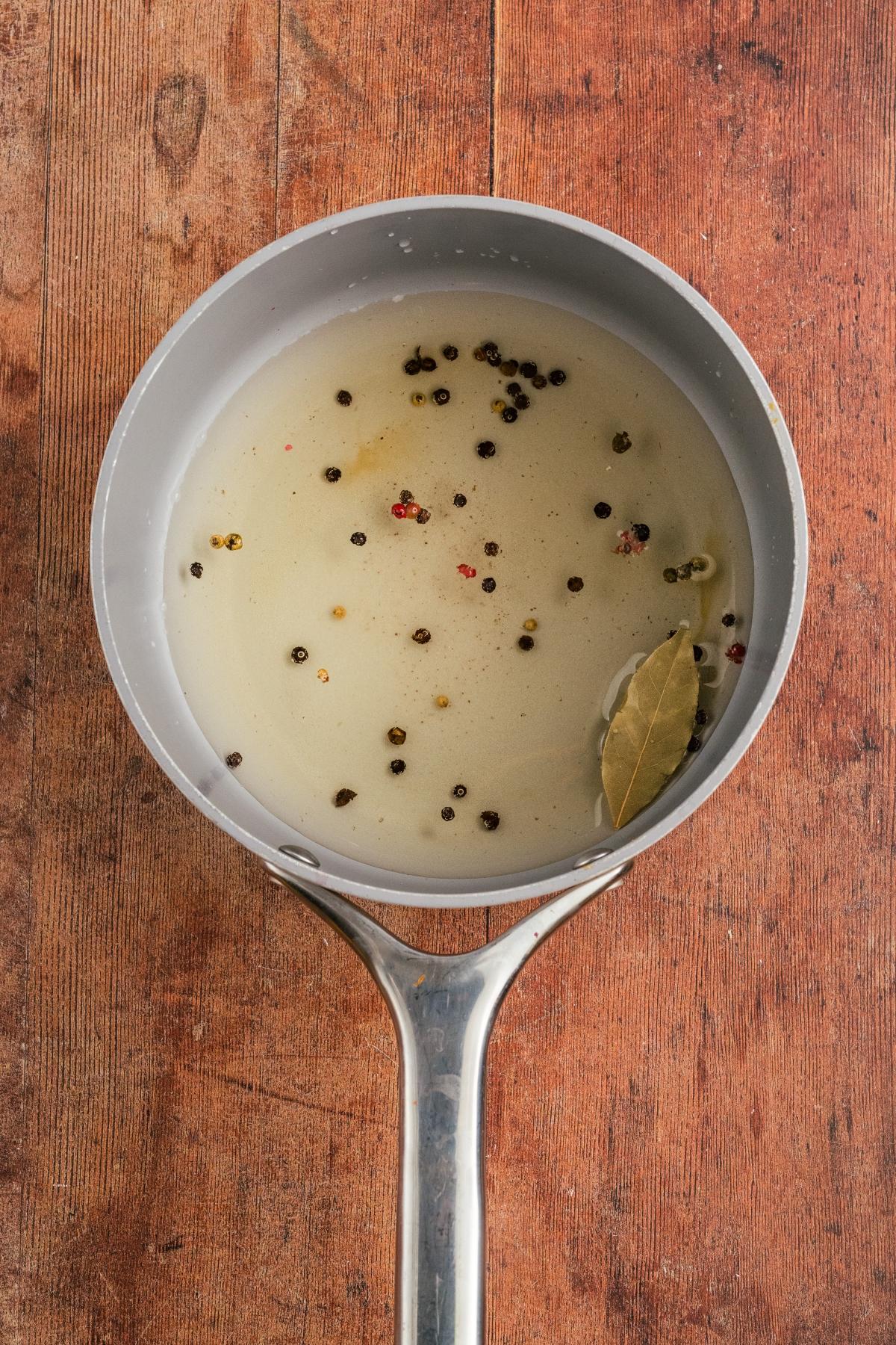 Gray saucepan with liquid, peppercorns, and a bay leaf on a wooden surface, viewed from above.