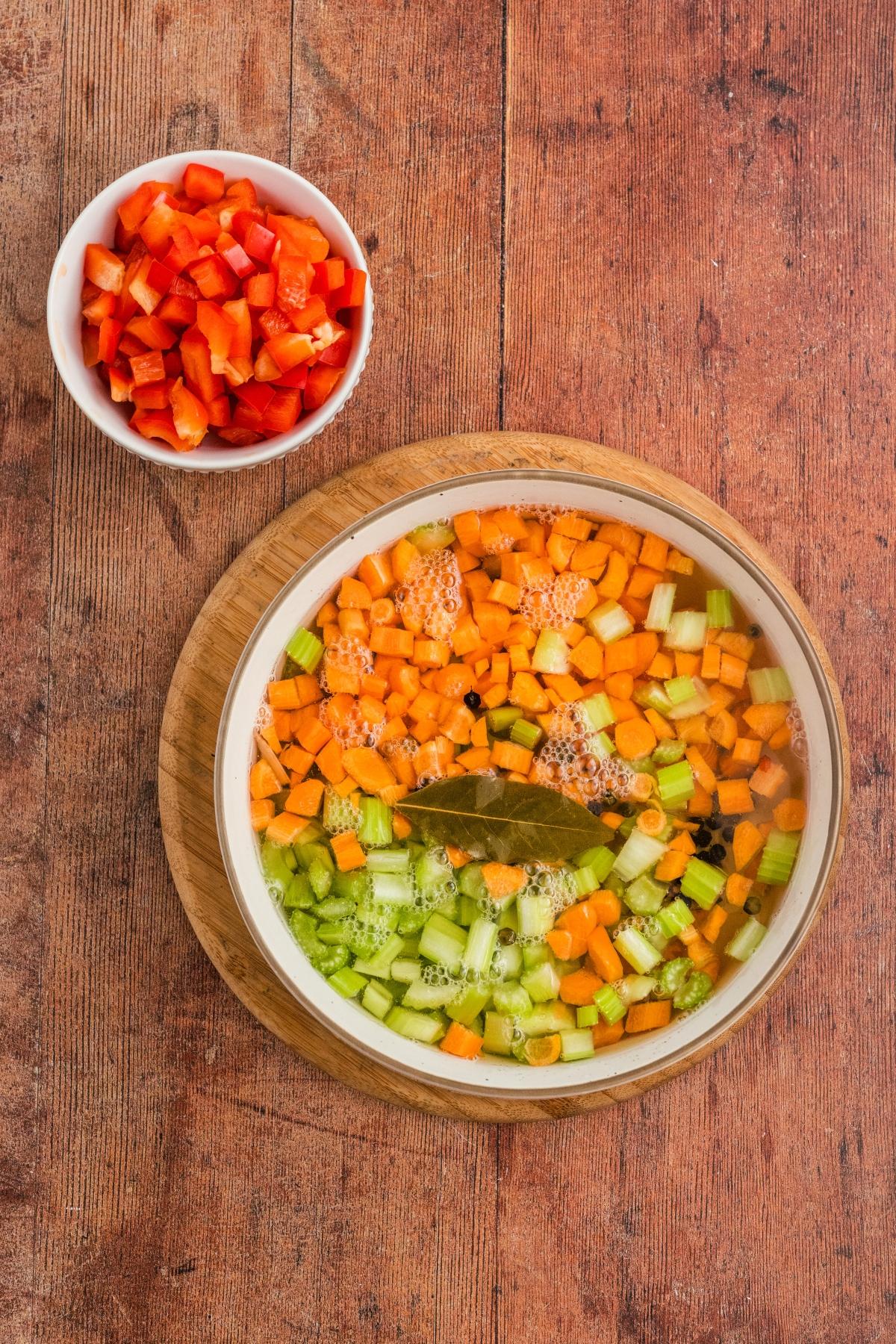 Chopped carrots, celery, and bay leaf simmering in a pot; diced red bell peppers in a bowl nearby.
