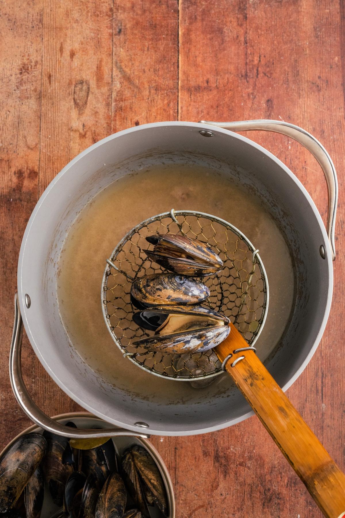 A slotted spoon lifts cooked mussels from a pot of broth on a wooden surface.