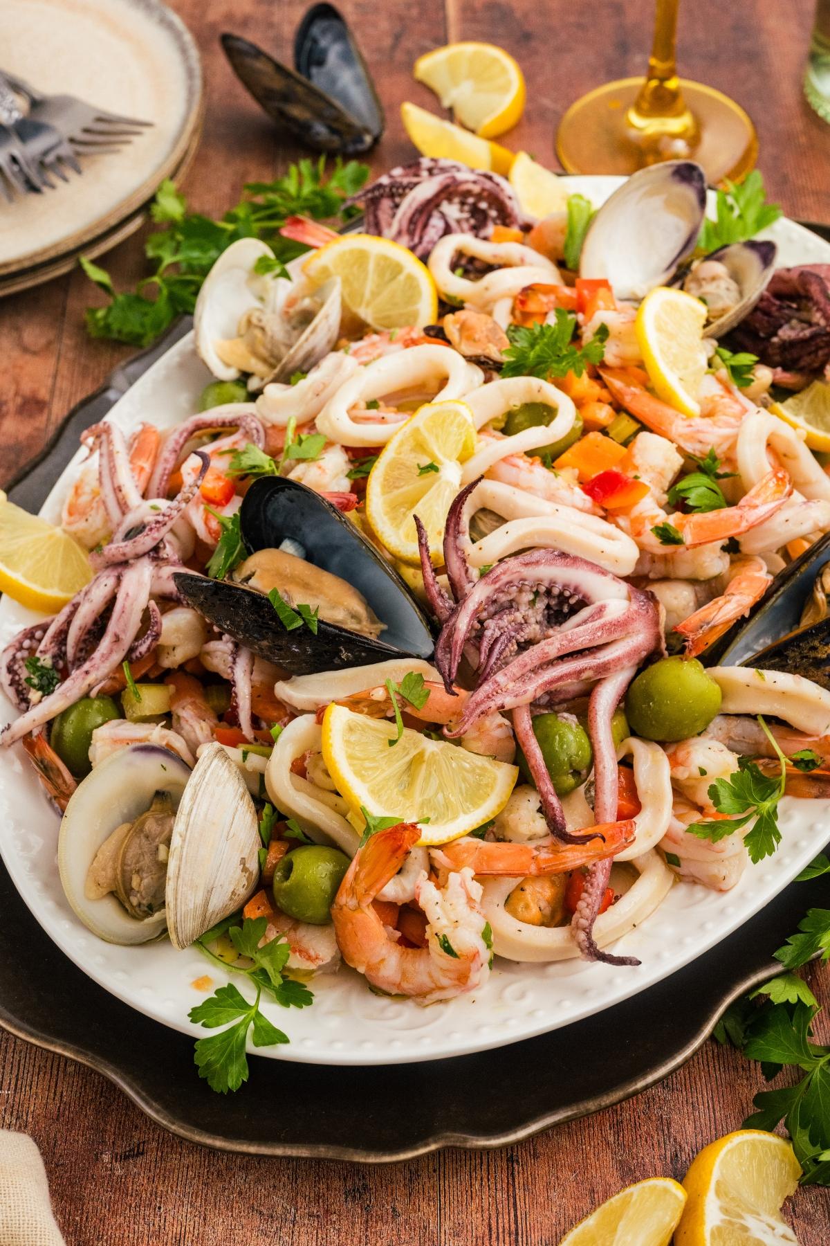 A platter of seafood salad with shrimp, squid, mussels, clams, olives, lemon slices, and fresh parsley.
