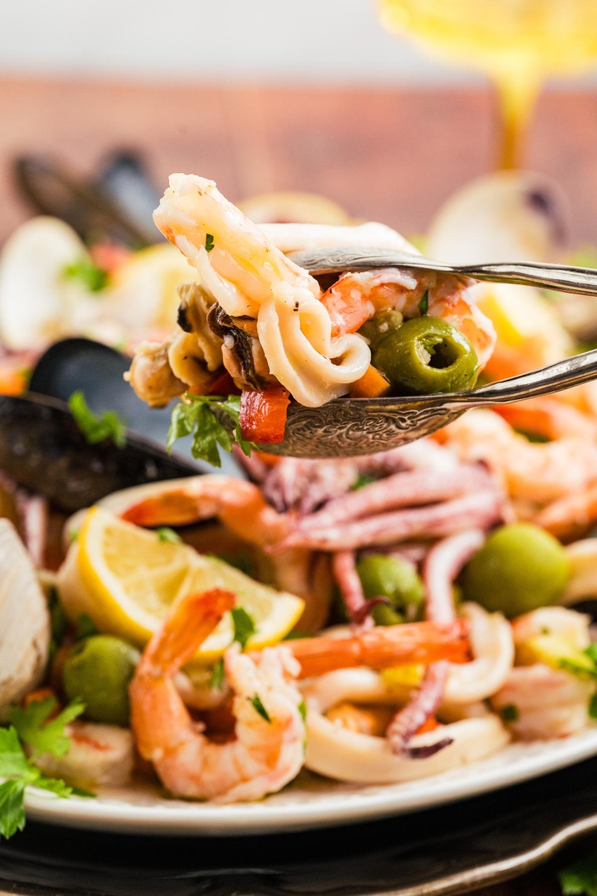 Tongs holding a serving of seafood salad with shrimp, calamari, olives, lemon, and parsley.