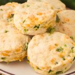 A plate of round Jalapeño Cheddar Biscuits, topped with visible cheese and green jalapeño pieces, sits on a woven placemat.