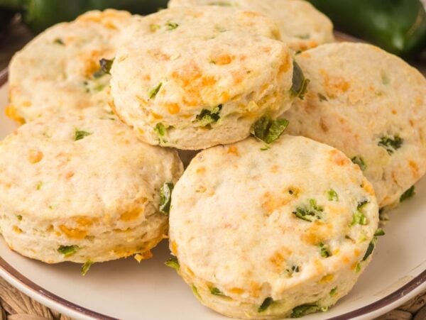 A plate of round Jalapeño Cheddar Biscuits, topped with visible cheese and green jalapeño pieces, sits on a woven placemat.
