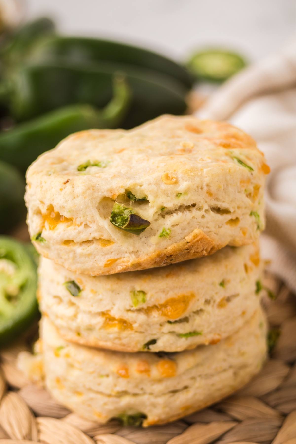 Three flaky biscuits with visible green and yellow pieces, stacked on a woven surface with jalapeños in background.