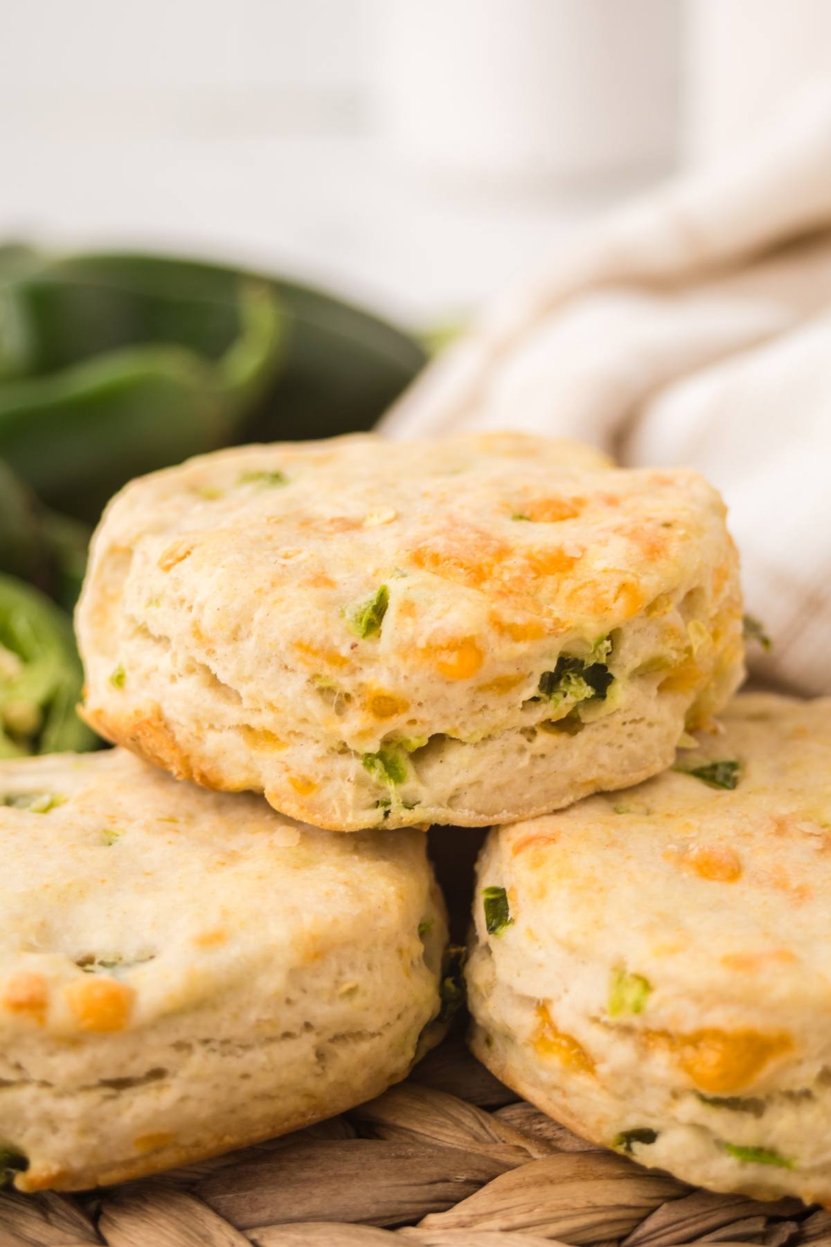 Three biscuits with visible bits of cheese and green peppers stacked on a woven mat, with peppers in background.