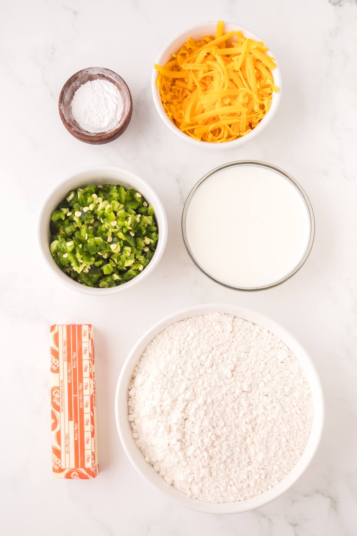 Ingredients arranged on a white surface: flour, cheddar cheese, milk, chopped jalapeños, butter, and baking powder.