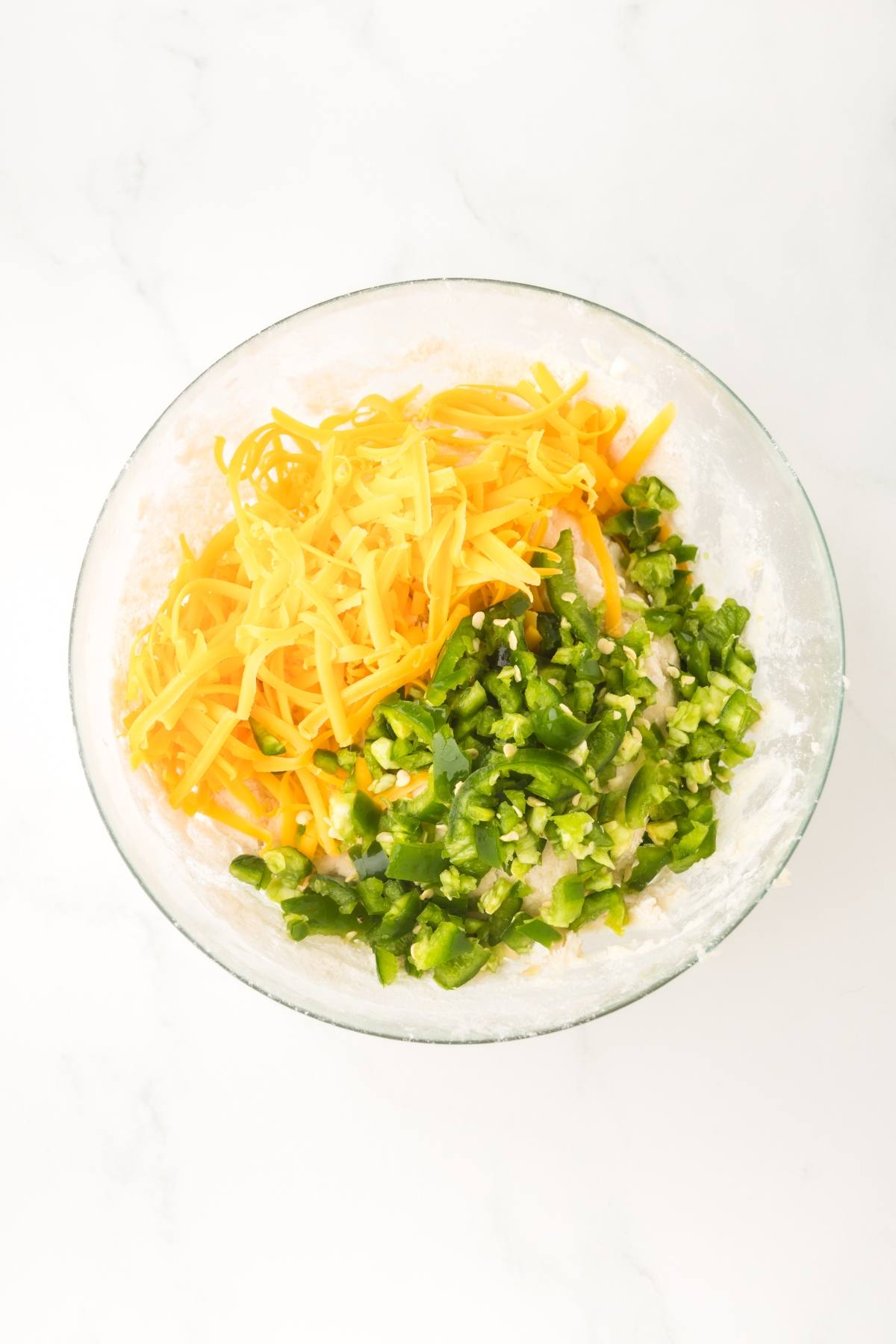 Clear bowl with shredded cheddar cheese and chopped green peppers on a flour mixture, seen from above.
