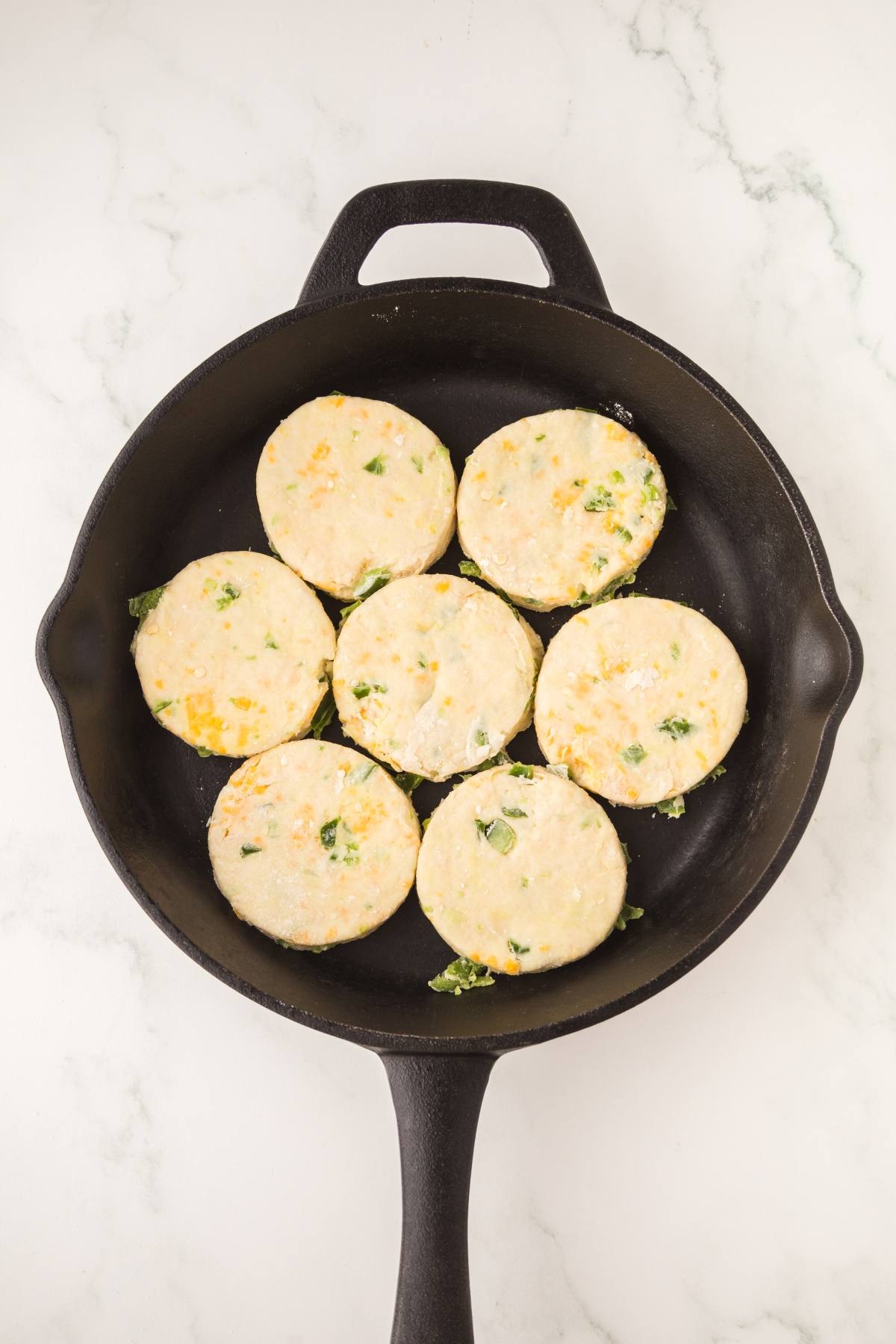 Seven uncooked round biscuits with herbs in a black skillet on a white marble surface.