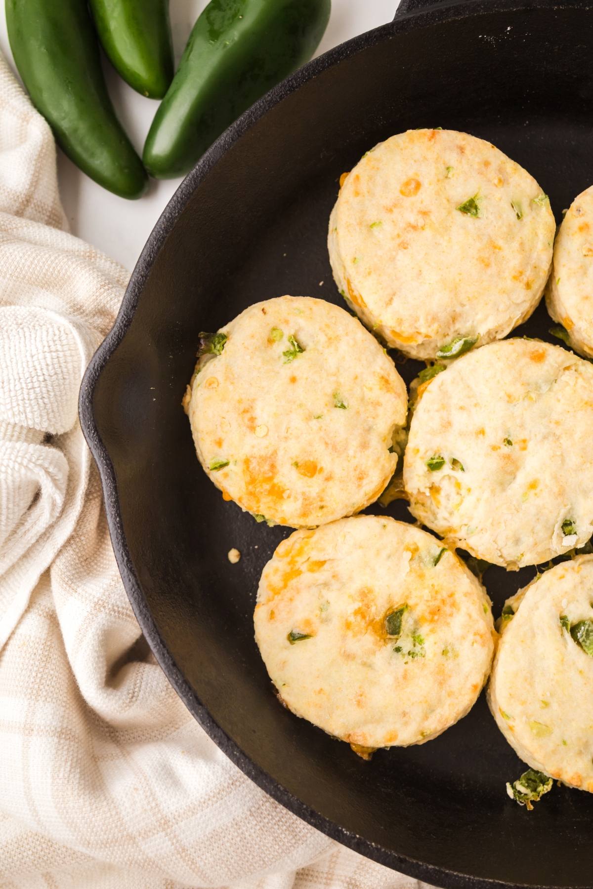 Six jalapeño biscuits in a cast iron skillet, with fresh jalapeños and a towel nearby.