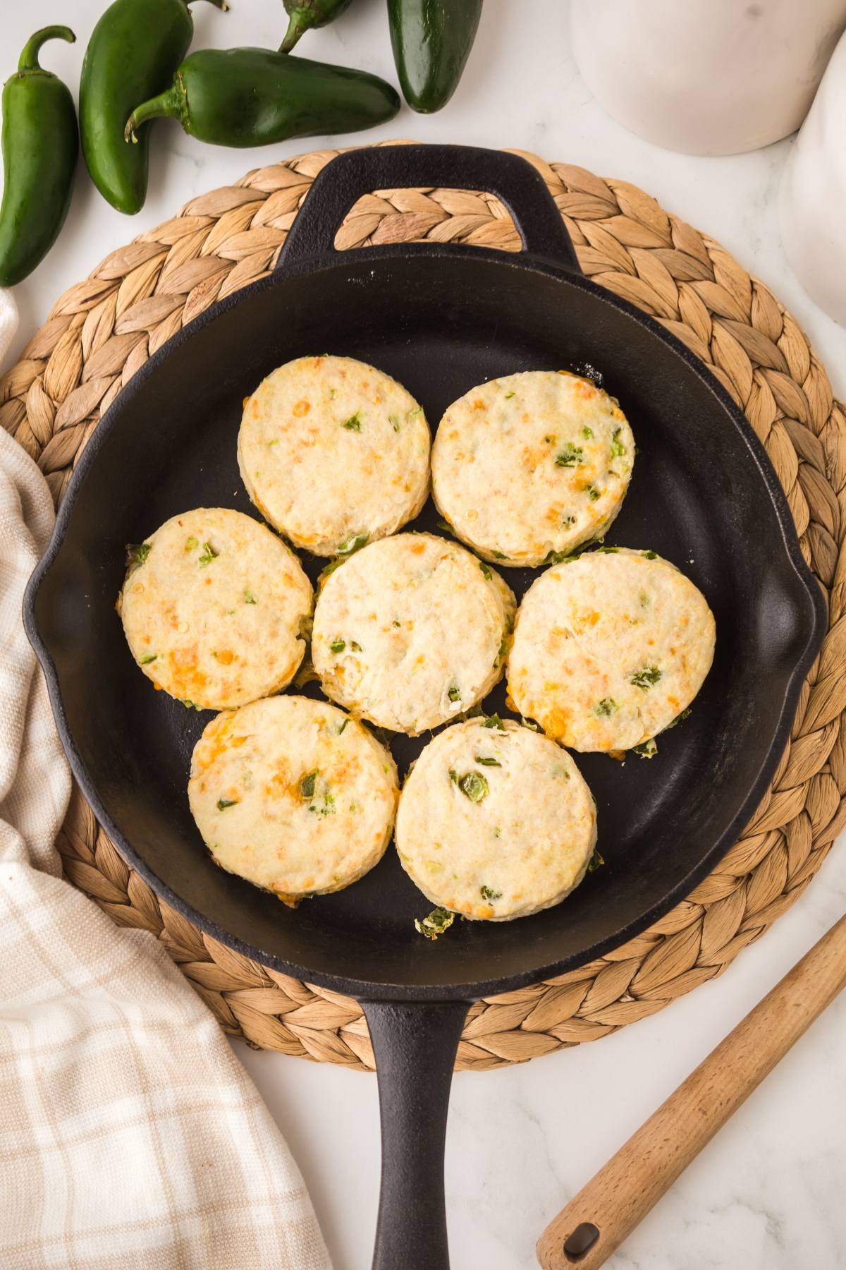Seven biscuits with green herbs in a black skillet on a woven mat, with jalapeños and a towel nearby.