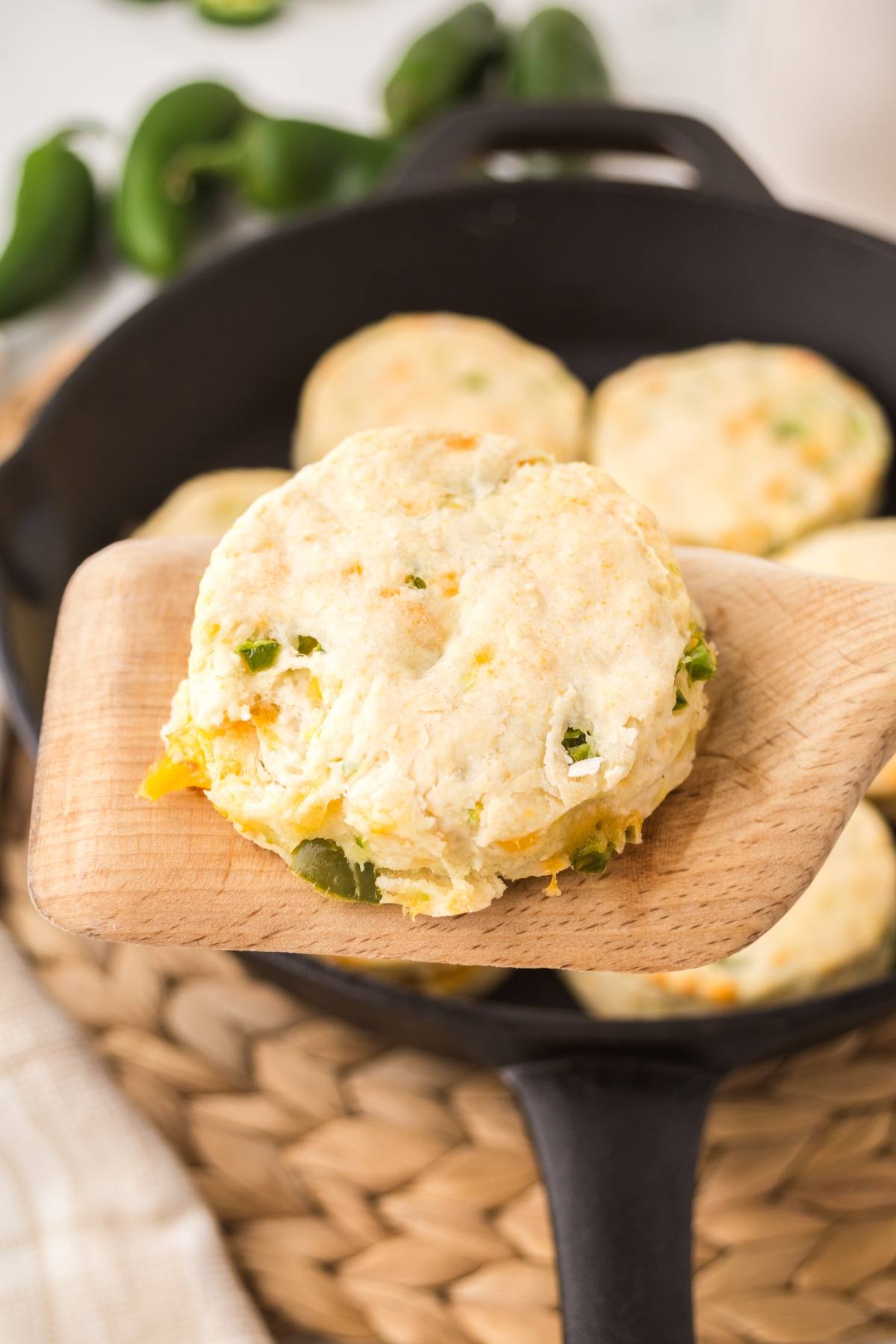A wooden spatula holds a cheesy biscuit with green bits over a skillet filled with more biscuits.