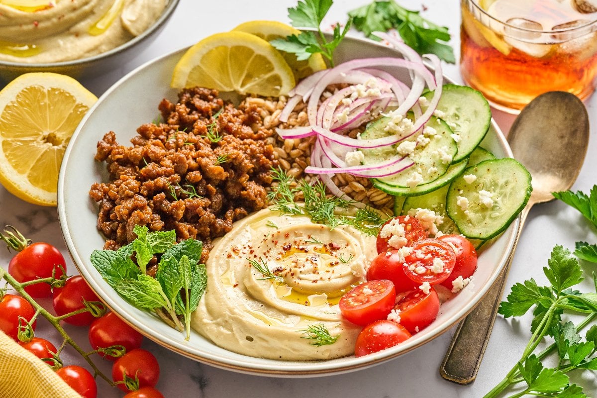 A bowl with lamb hummus, spiced ground meat, cucumber, tomatoes, red onion, lemon slices, and fresh herbs.