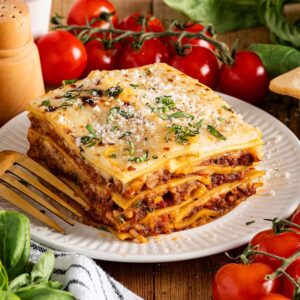 A slice of lasagna alla Bolognese on a plate with fresh tomatoes, basil, parmesan cheese, and a fork beside it.