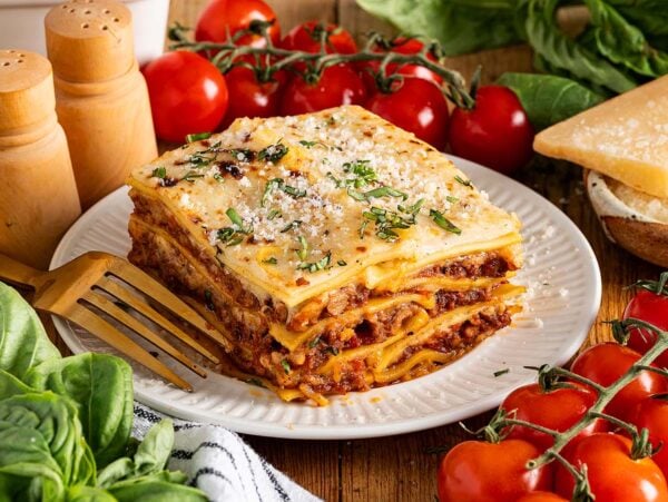 A slice of lasagna alla Bolognese on a plate with fresh tomatoes, basil, parmesan cheese, and a fork beside it.
