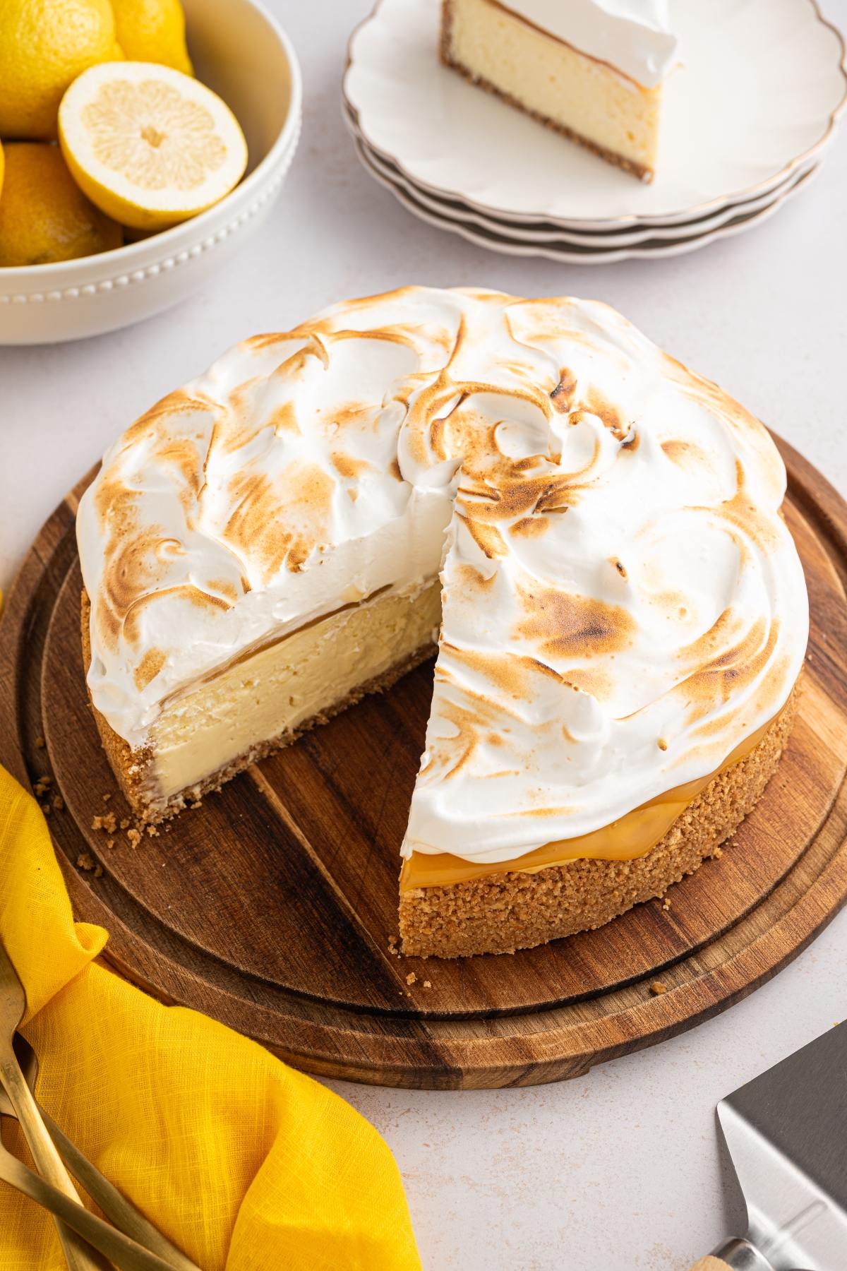 Lemon meringue cheesecake on a wooden board with one slice removed, surrounded by lemons and plates.