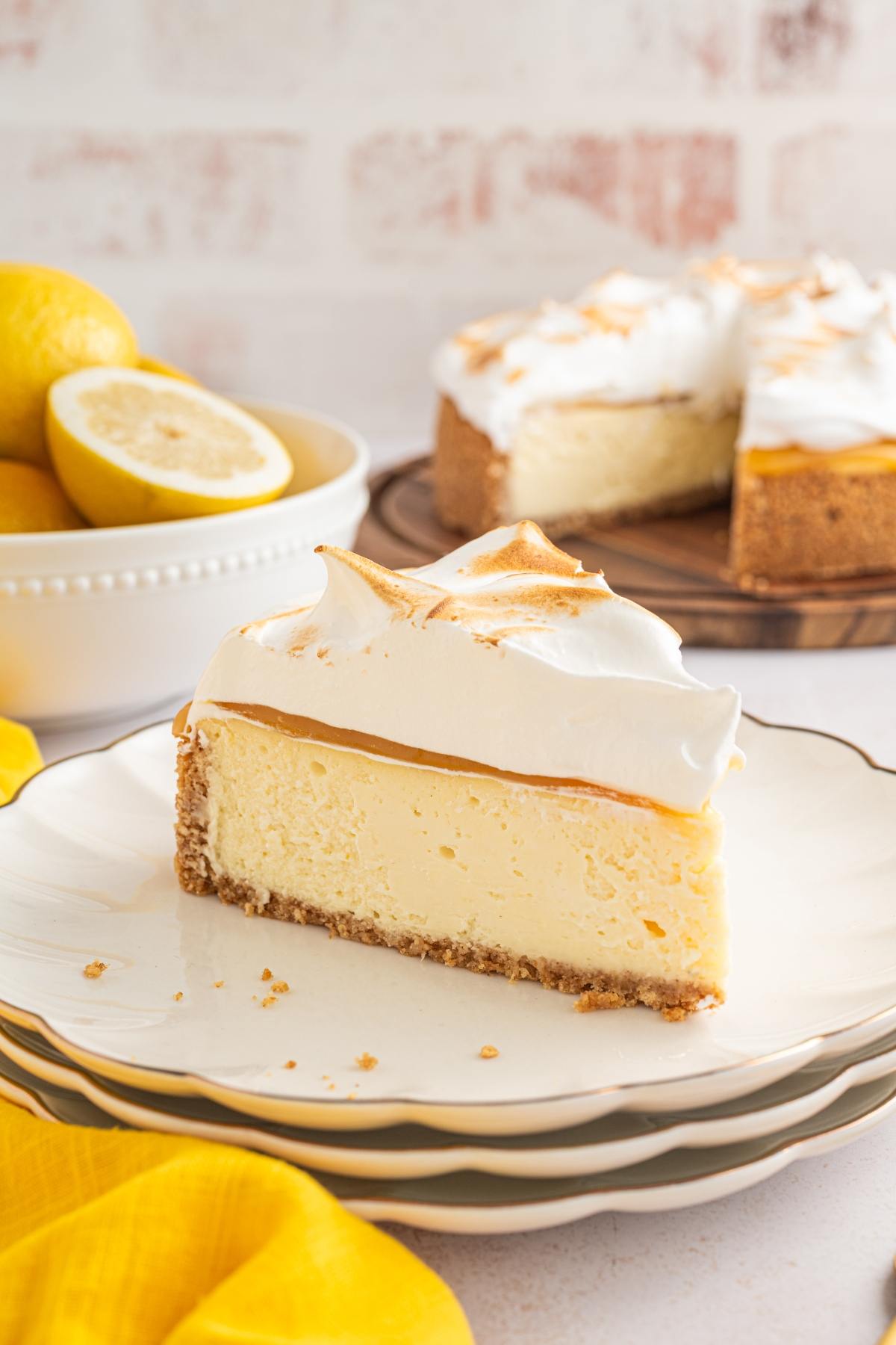 A slice of lemon meringue cheesecake on a plate, with whole and cut lemons in the background.