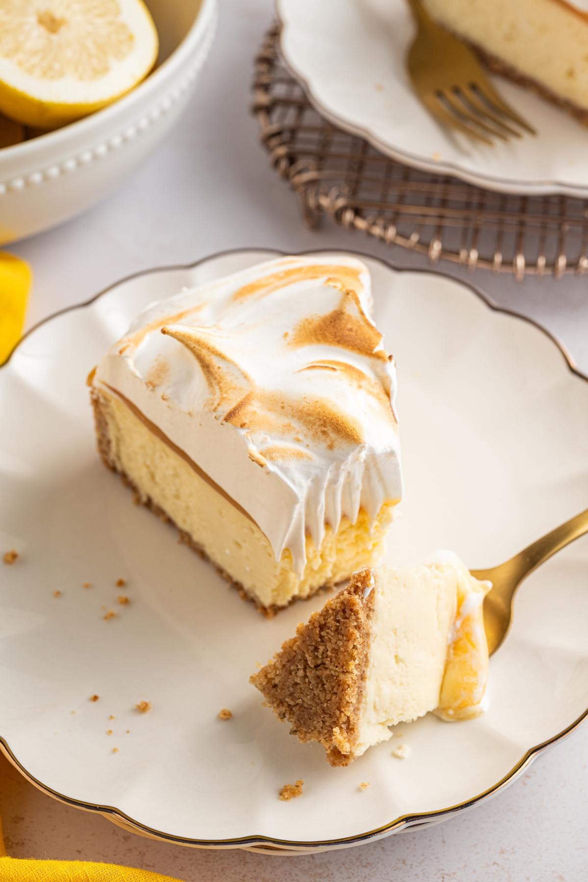 A slice of lemon meringue cheesecake on a plate with a forkful cut out, next to a bowl of lemons.