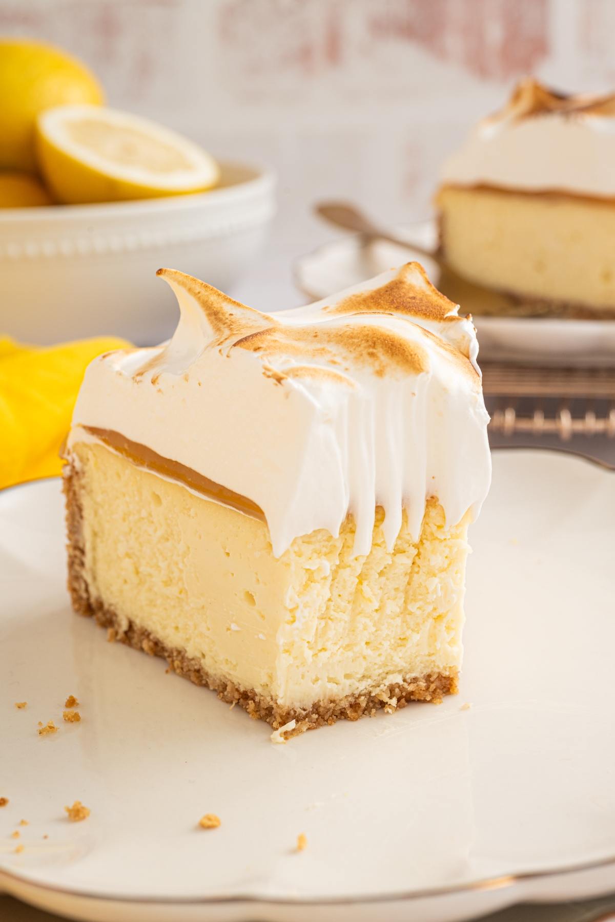 A slice of lemon meringue cheesecake on a plate, with a bowl of lemons in the background.