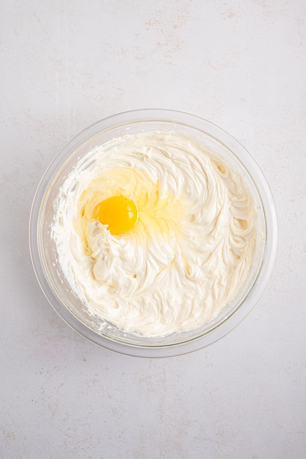 A glass bowl of whipped cream cheese mixture with a raw egg on top.