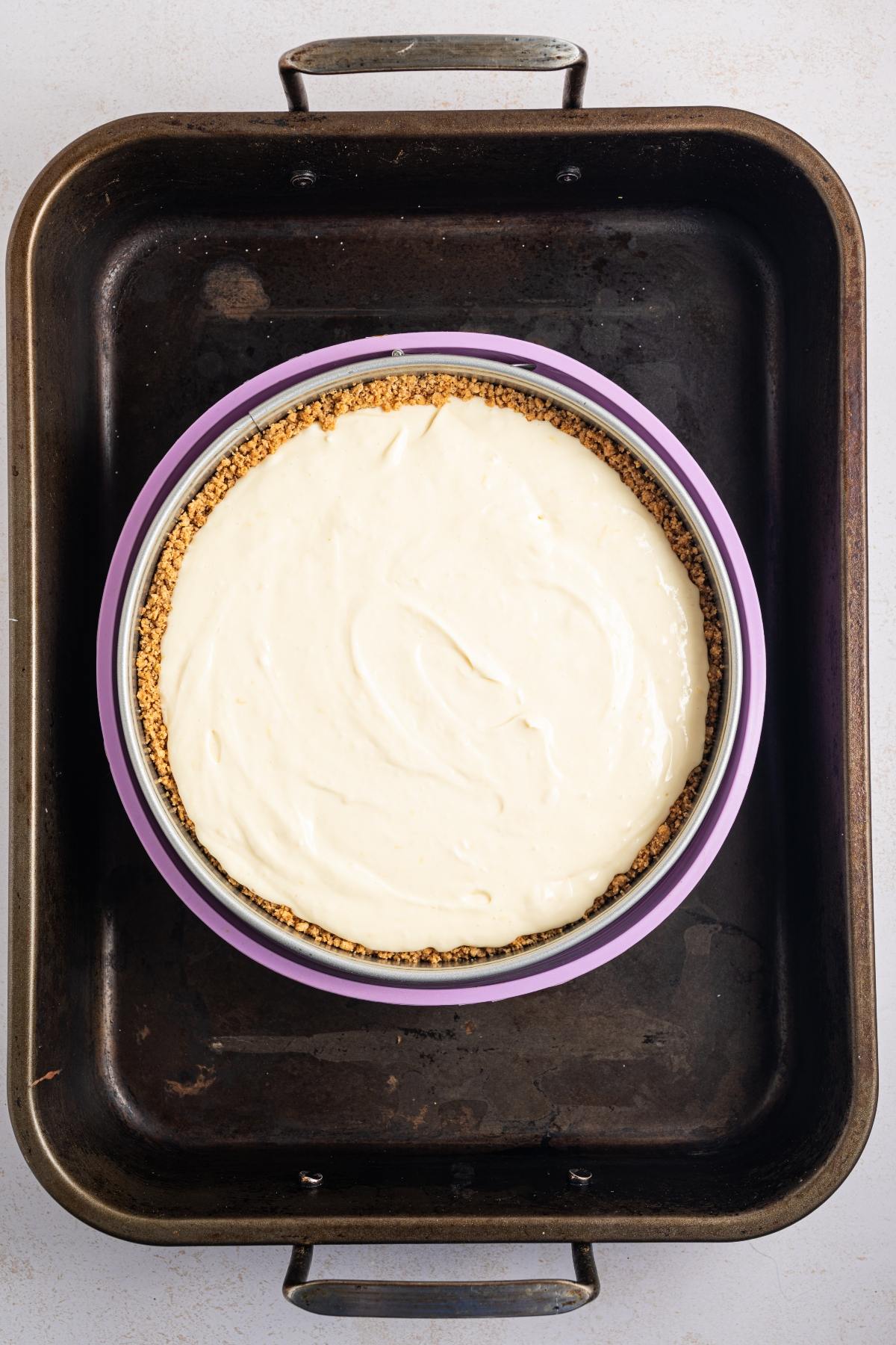 A cheesecake in a springform pan sits in a large roasting pan, ready to be baked.