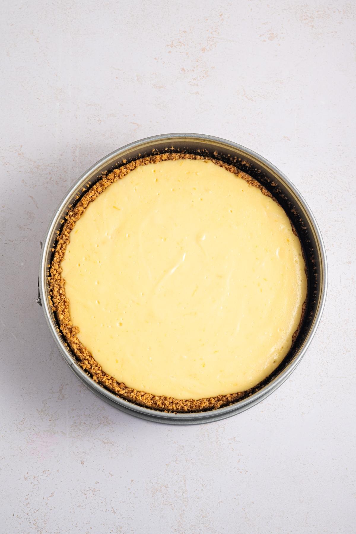 A round cheesecake with a golden graham cracker crust in a springform pan on a light surface.