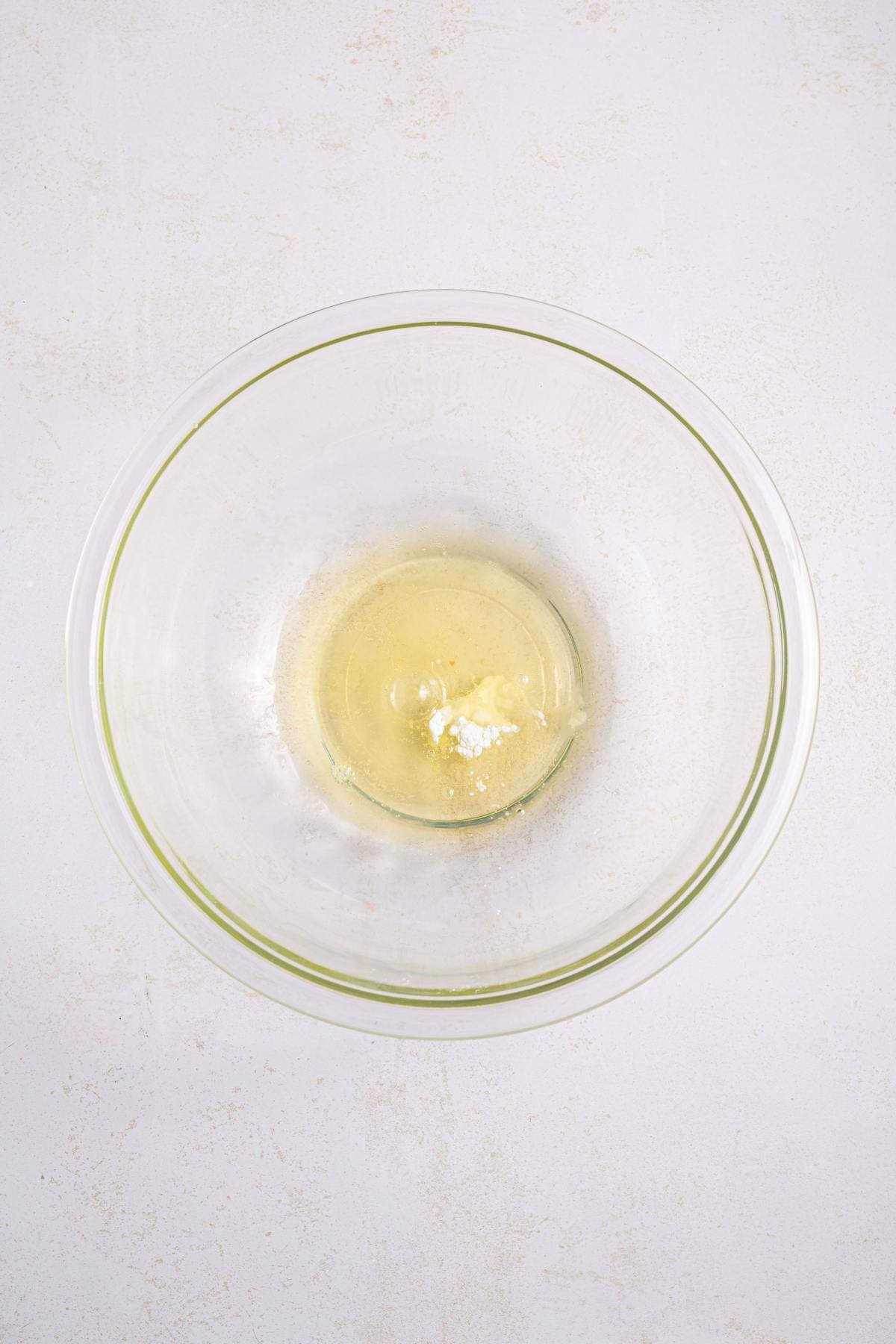 A glass bowl with egg whites and a pinch of salt on a light-colored surface.