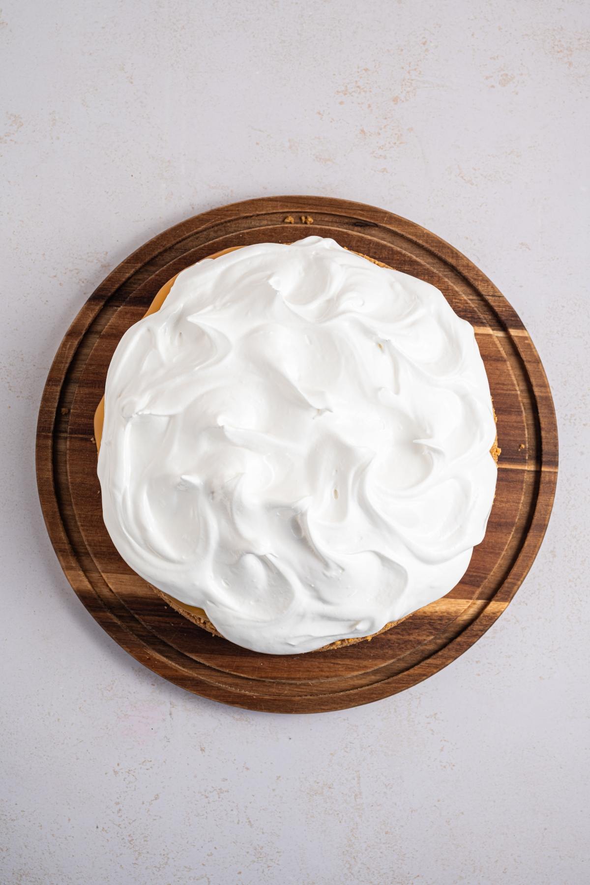 A round cake topped with swirled white meringue sits on a wooden board against a light background.