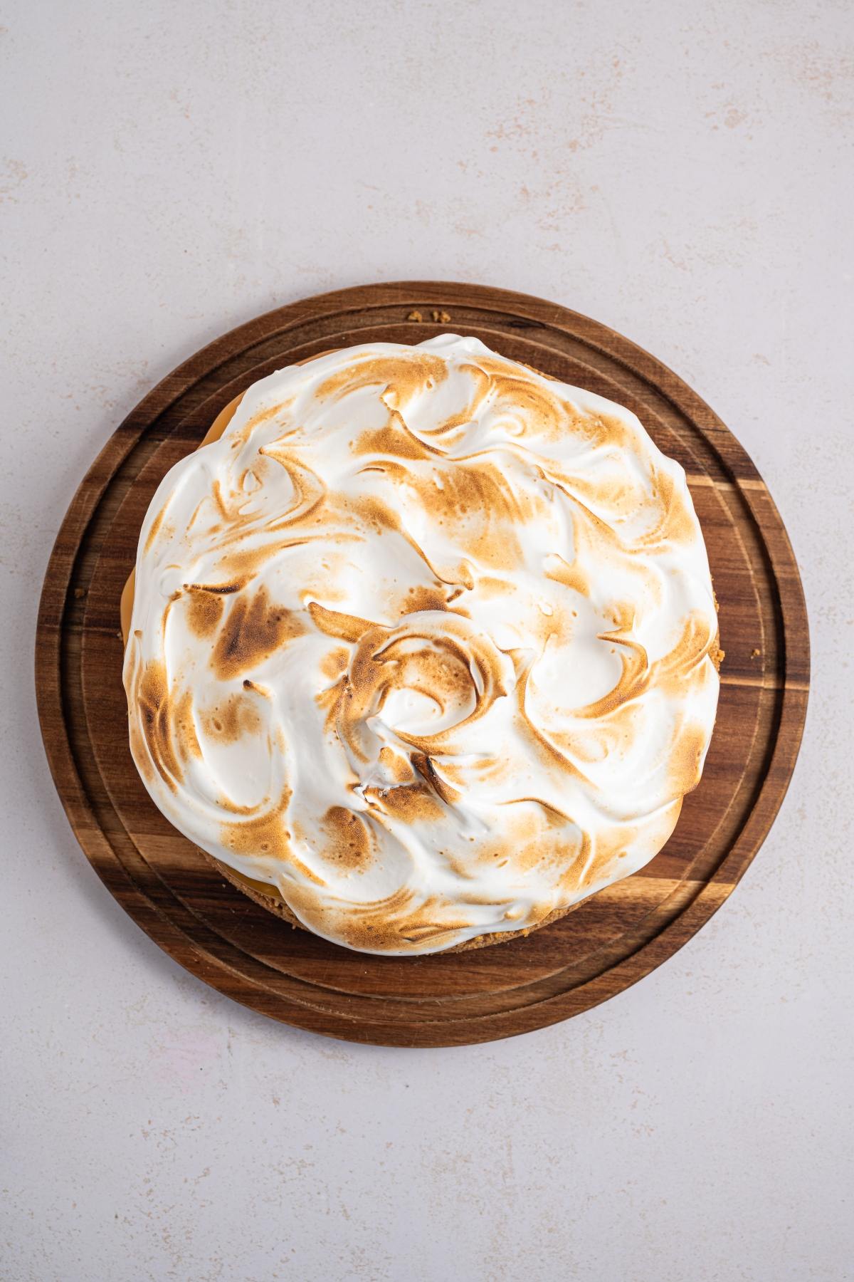 A round cake with toasted meringue topping on a wooden board, viewed from above.