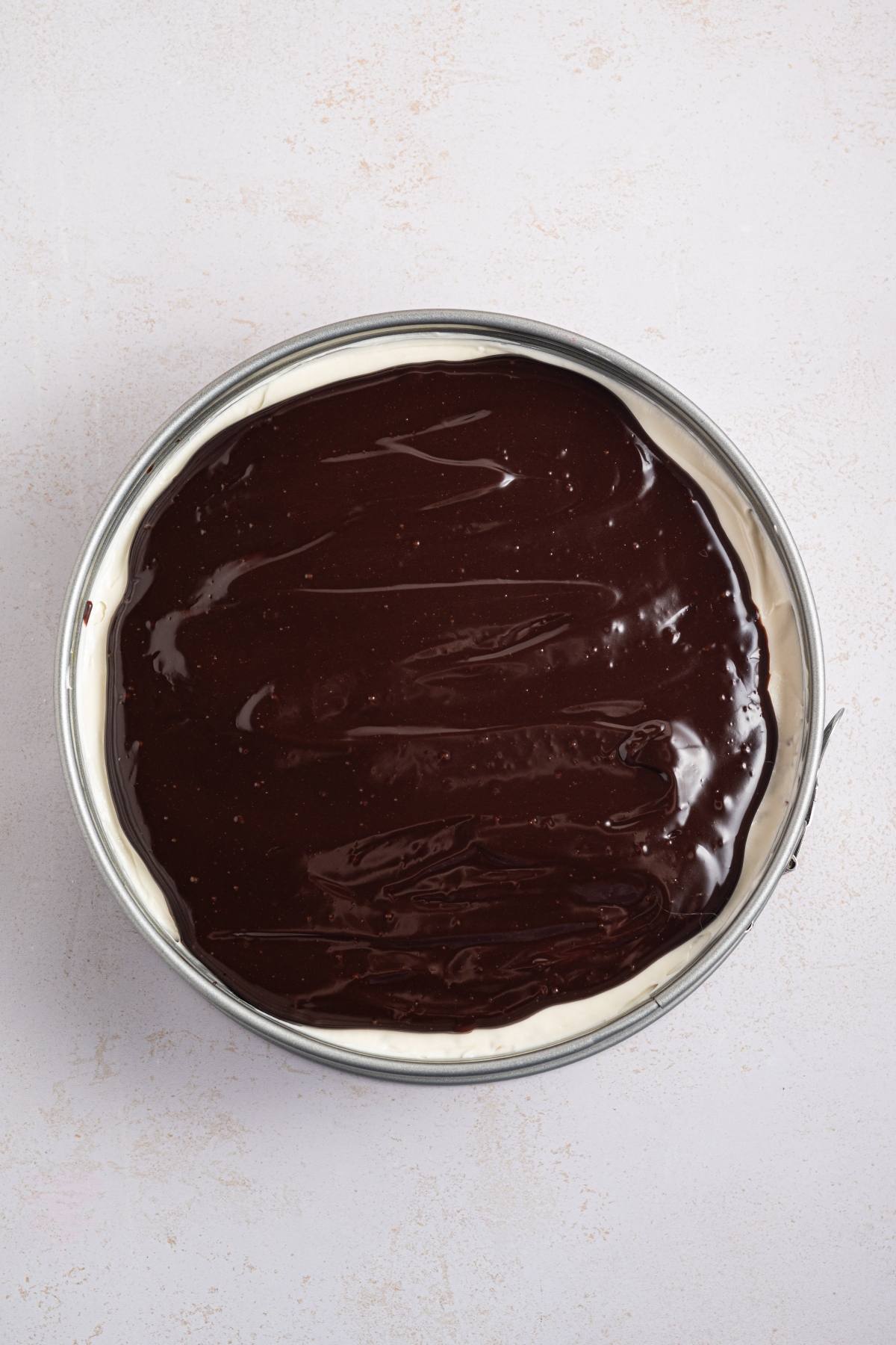 A round cheesecake topped with a smooth, glossy layer of chocolate ganache in a metal pan.