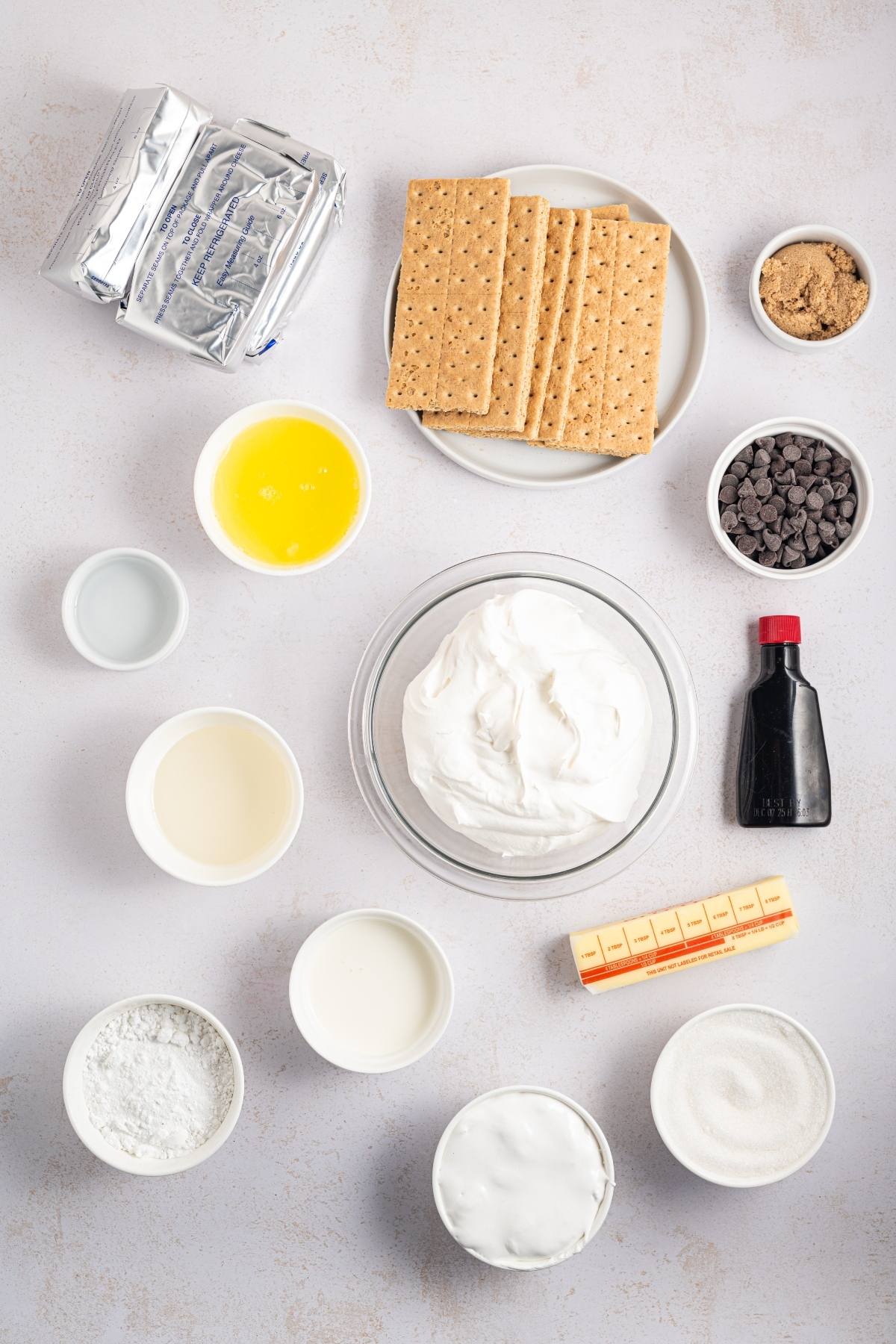 Top-down view of s'mores cheesecake ingredients including graham crackers, chocolate chips, and butter on a white surface.