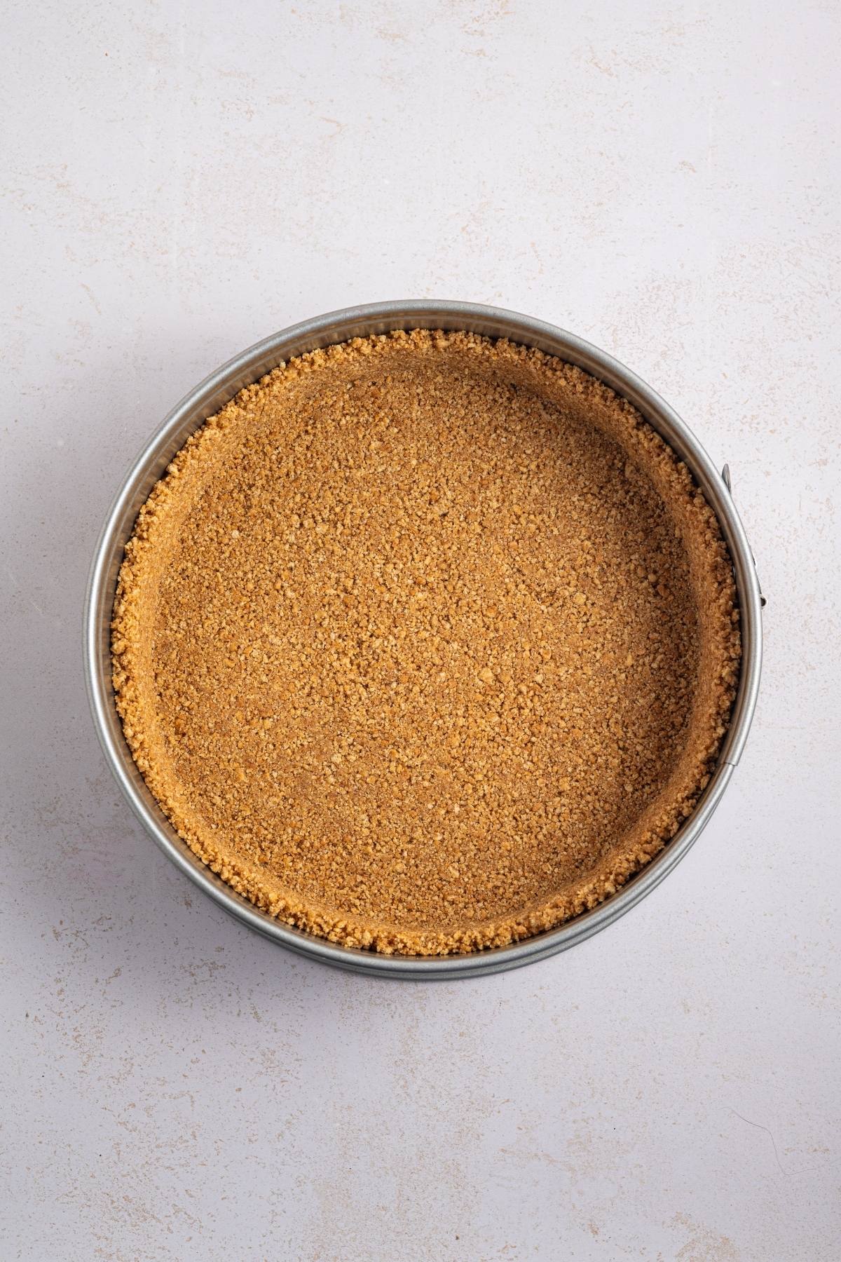 A round pan filled with a pressed graham cracker crust on a light-colored surface.