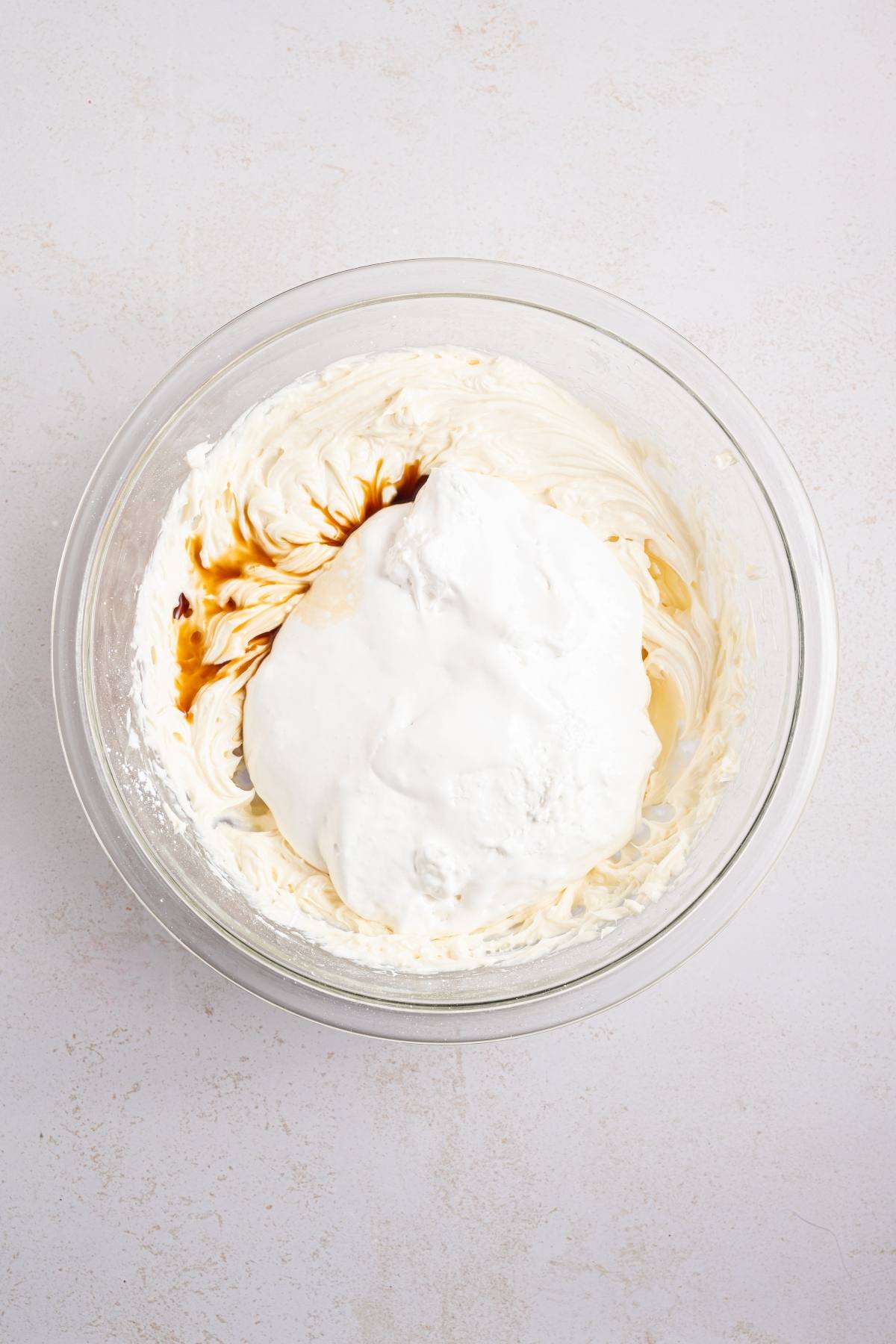 A glass bowl with whipped cream, vanilla extract, and marshmallow fluff on a light surface.
