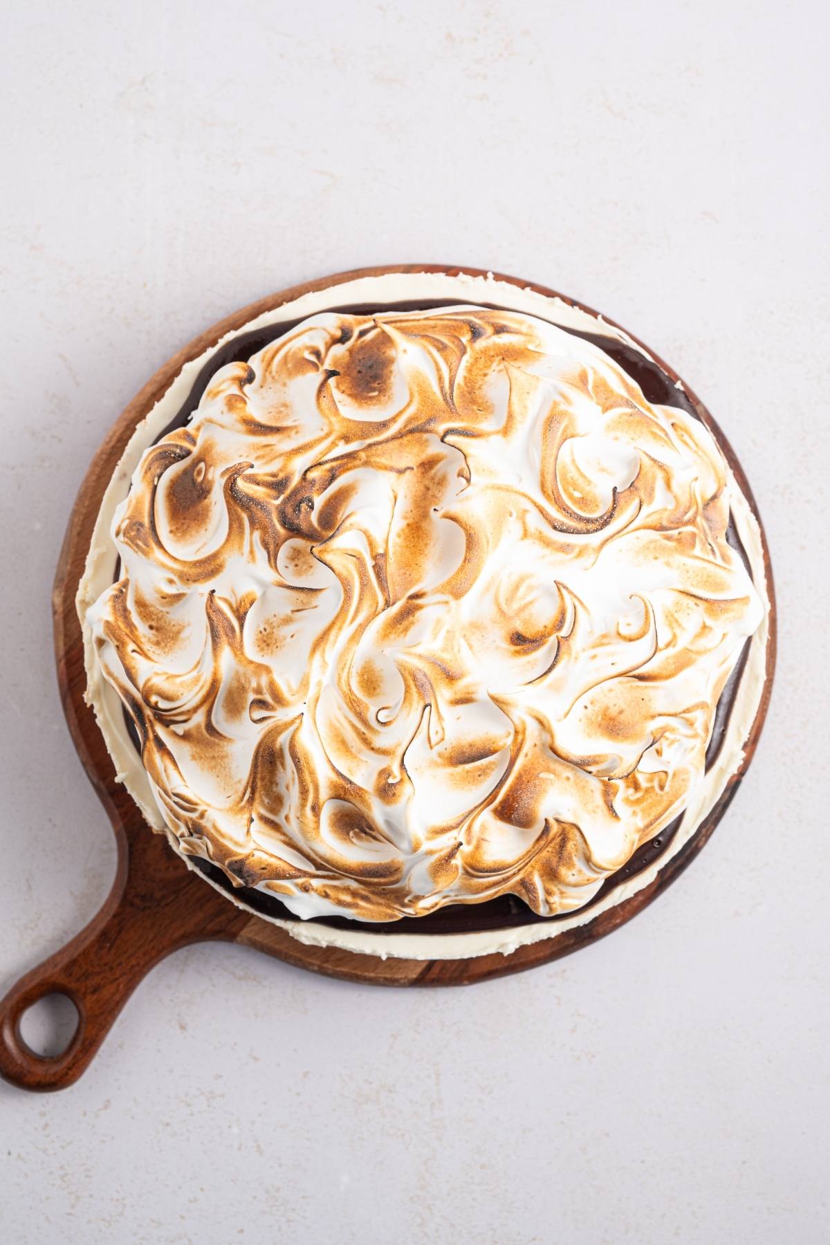 A round cake topped with toasted, swirled meringue on a wooden serving board.