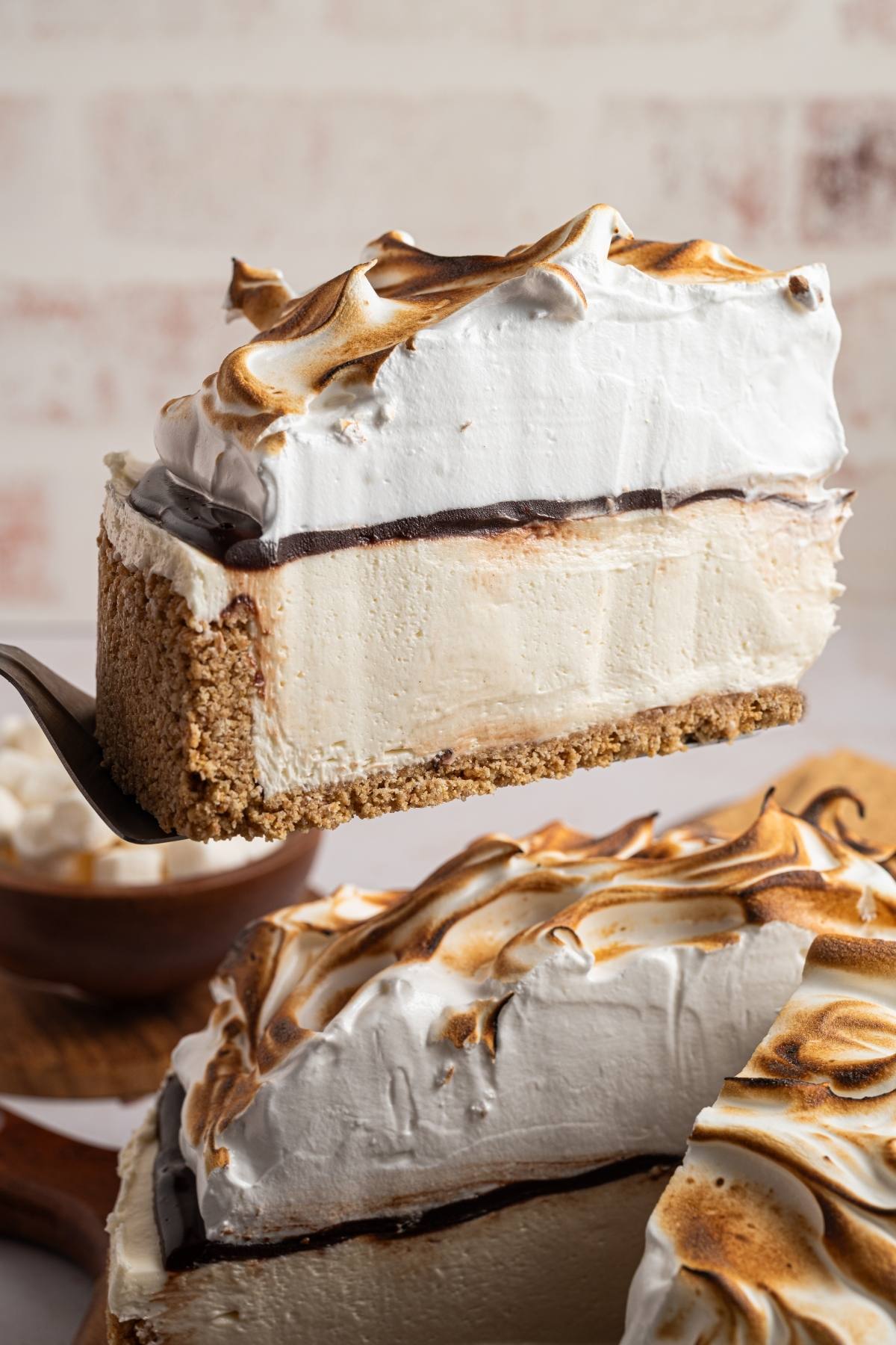 A slice of pie with graham crust, creamy filling, chocolate, and toasted meringue is lifted from the whole pie.