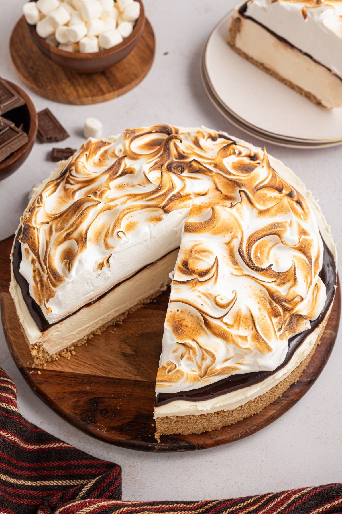 A s’mores cheesecake topped with toasted meringue, with one slice cut and set on a plate nearby.